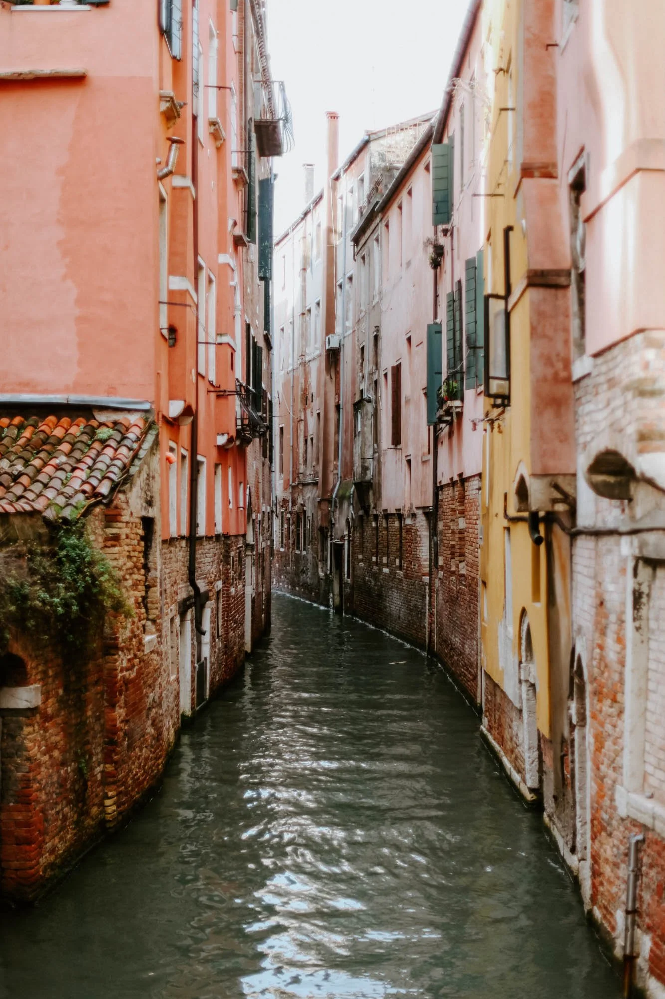 A narrow canal in Venice, Italy, lined with old, colorful buildings with green shutters and small windows, reflecting the historic European architecture.