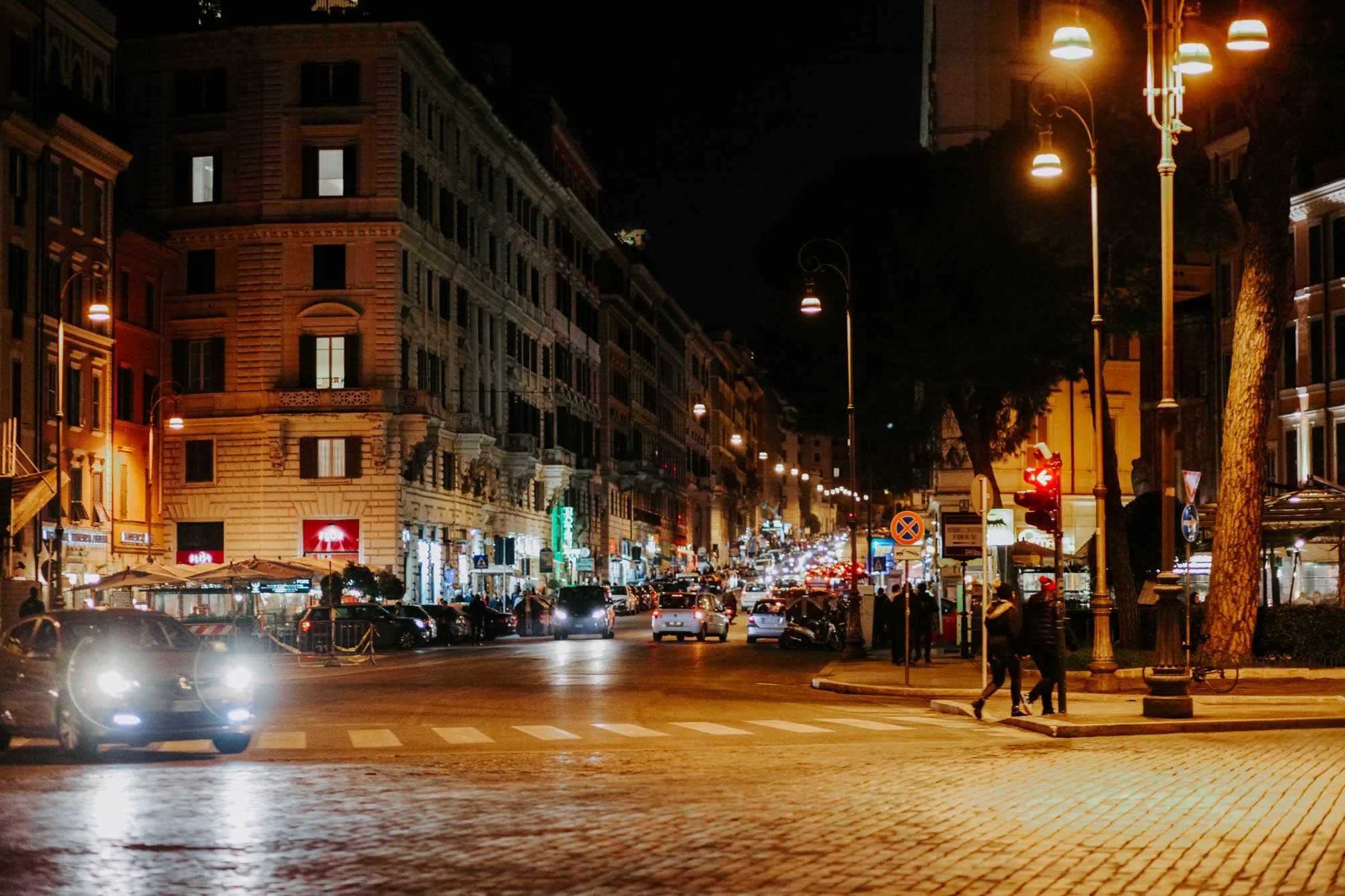 Night Rome street scene with cars, pedestrians, and illuminated buildings and streetlights.