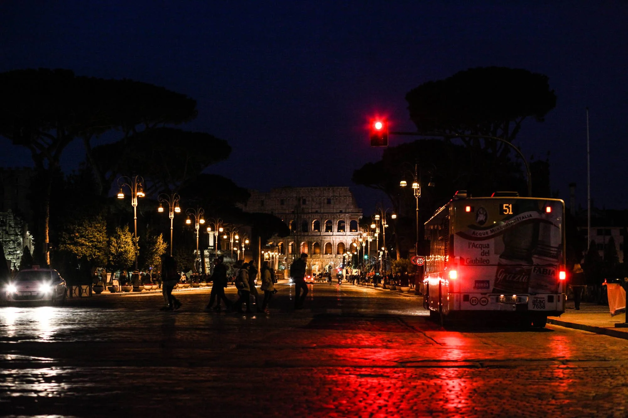 Nighttime scene in Rome with illuminated street lamps, people crossing at the pedestrian crosswalk, a bus with red taillights, and the Colosseum in the background under a dark sky.