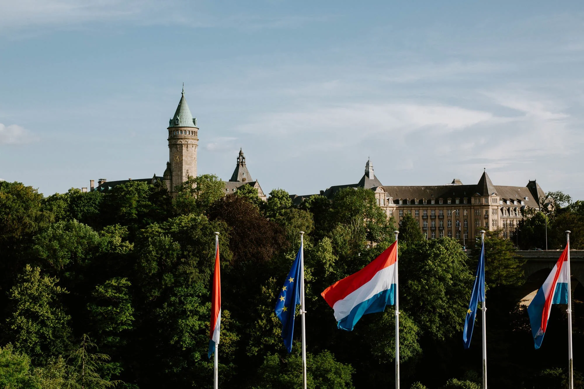 A historic castle-like building with a tall tower surrounded by green trees, with flags of the Netherlands, the European Union, and Luxembourg in the foreground.