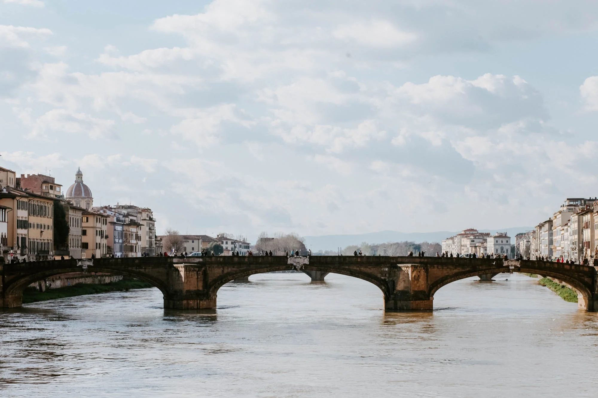 A river flows under an old stone bridge with arches, flanked by European-style buildings on both sides, with a church dome visible in the background, under a cloudy sky.
