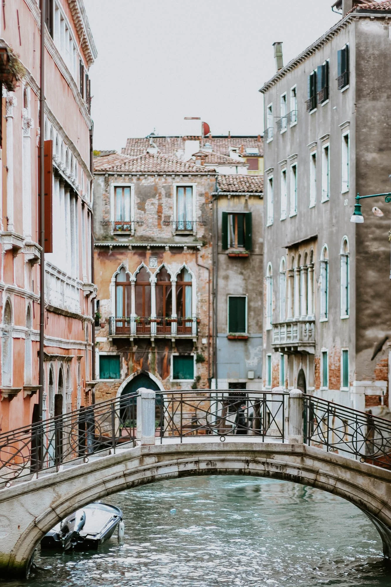 A canal in Venice, Italy, with a small bridge crossing over the water and historical buildings with arched windows and shutters lining the canal.