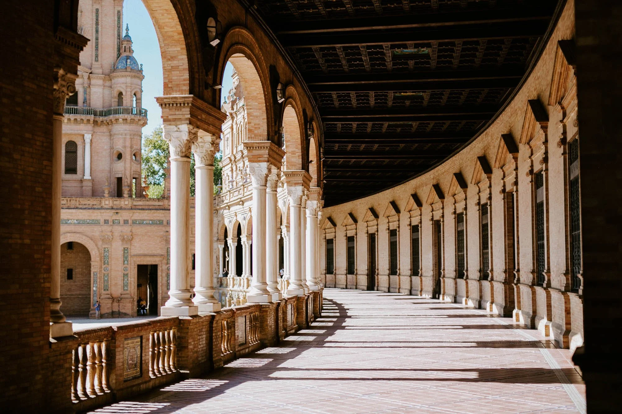 Arcaded walkway with brick and white columns, and a building with domes and spires seen in the background, on a sunny day in Seville, Spain