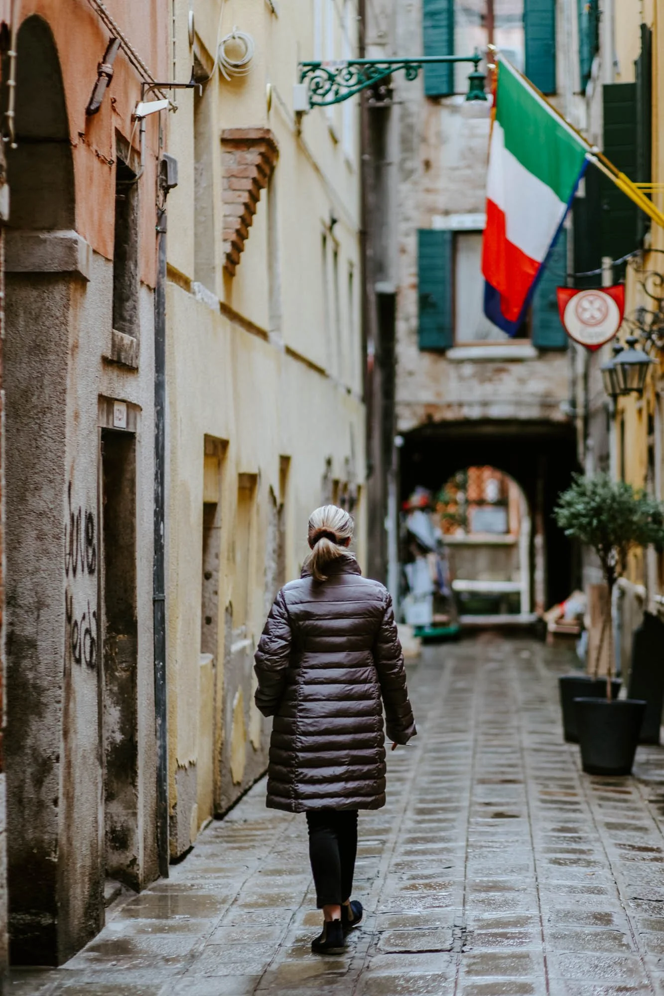 A woman walking down a wet, narrow street with old buildings, a flag of Italy, and potted plants.