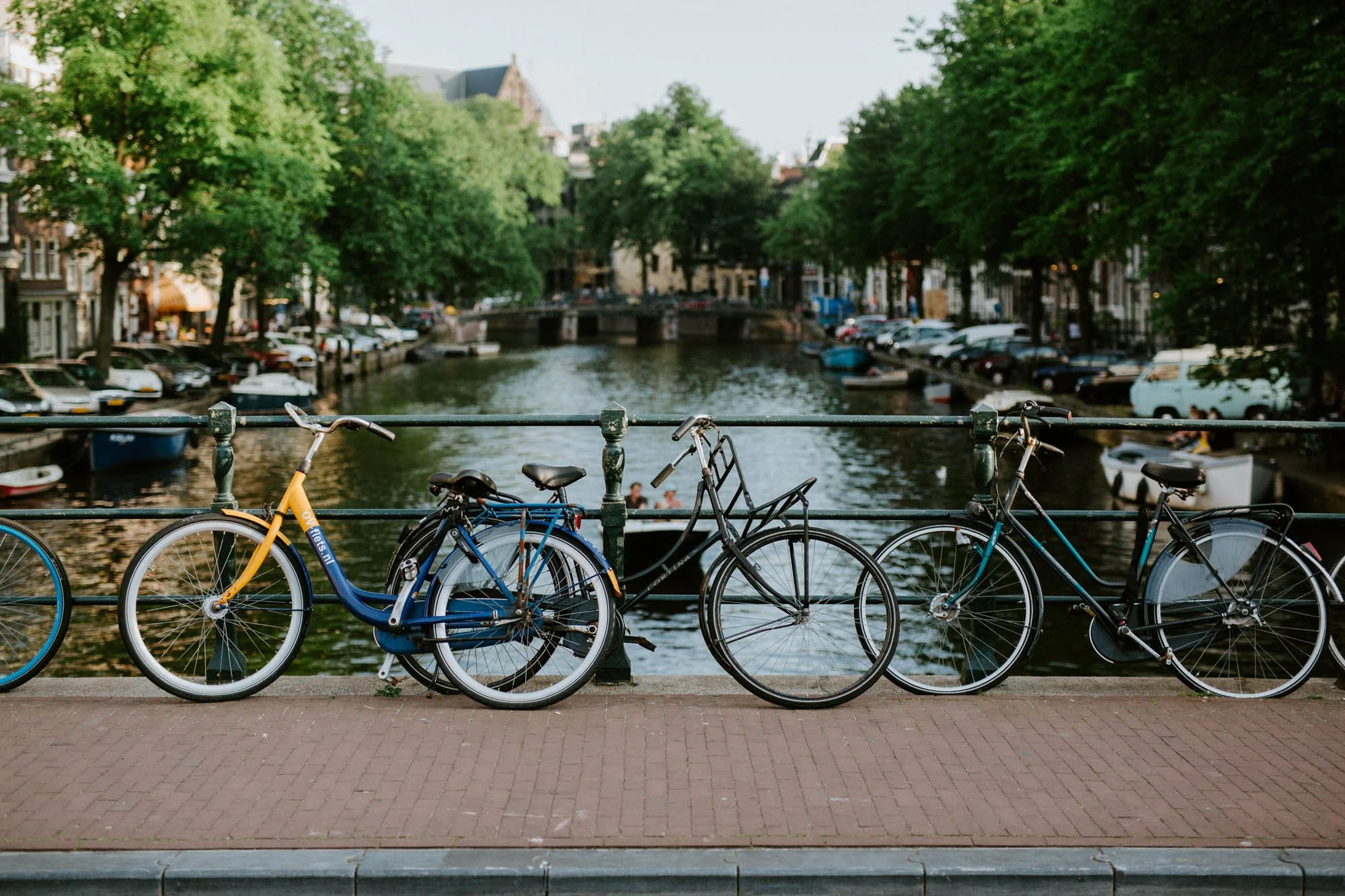 Three bicycles parked along a canal with trees, buildings, and boats in the background in Amsterdam.