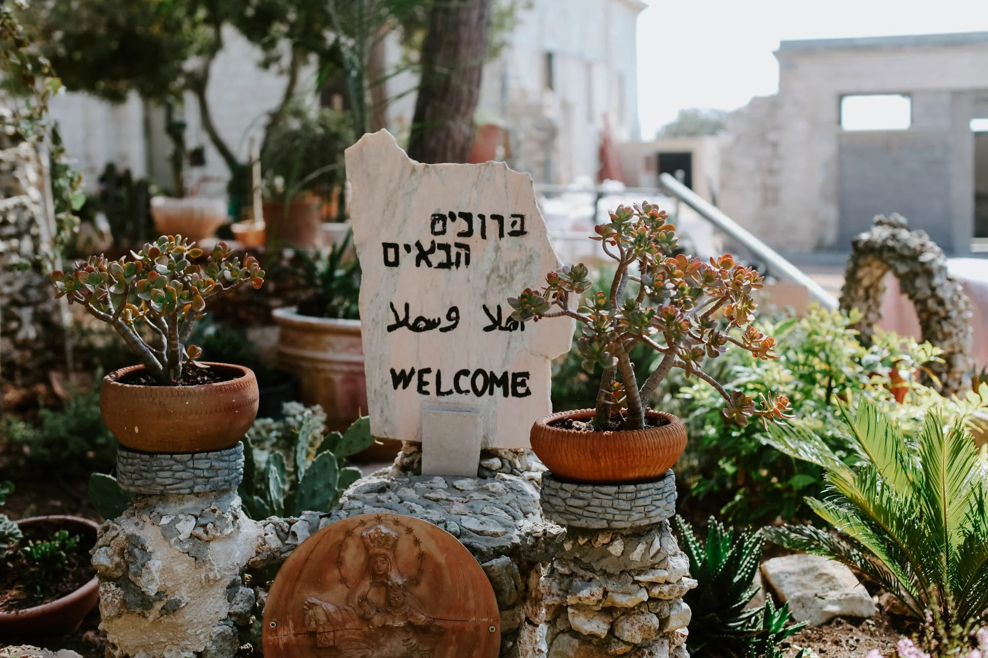 Garden with potted succulents and a white stone sign that says "Welcome" in multiple languages. There are trees and plants in the background in Nazareth.
