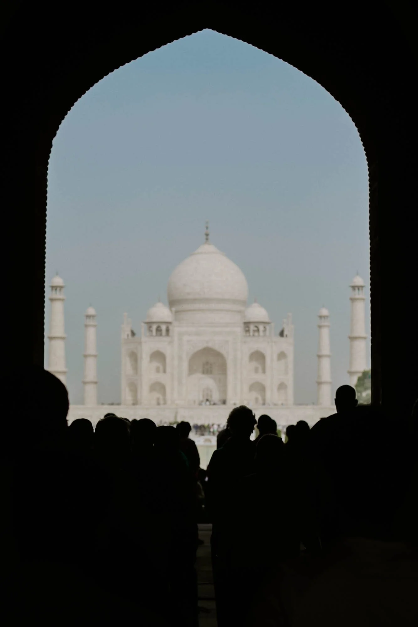 People silhouetted in a dark archway viewing the Taj Mahal in India, with the monument's white marble dome and minarets visible in the distance.