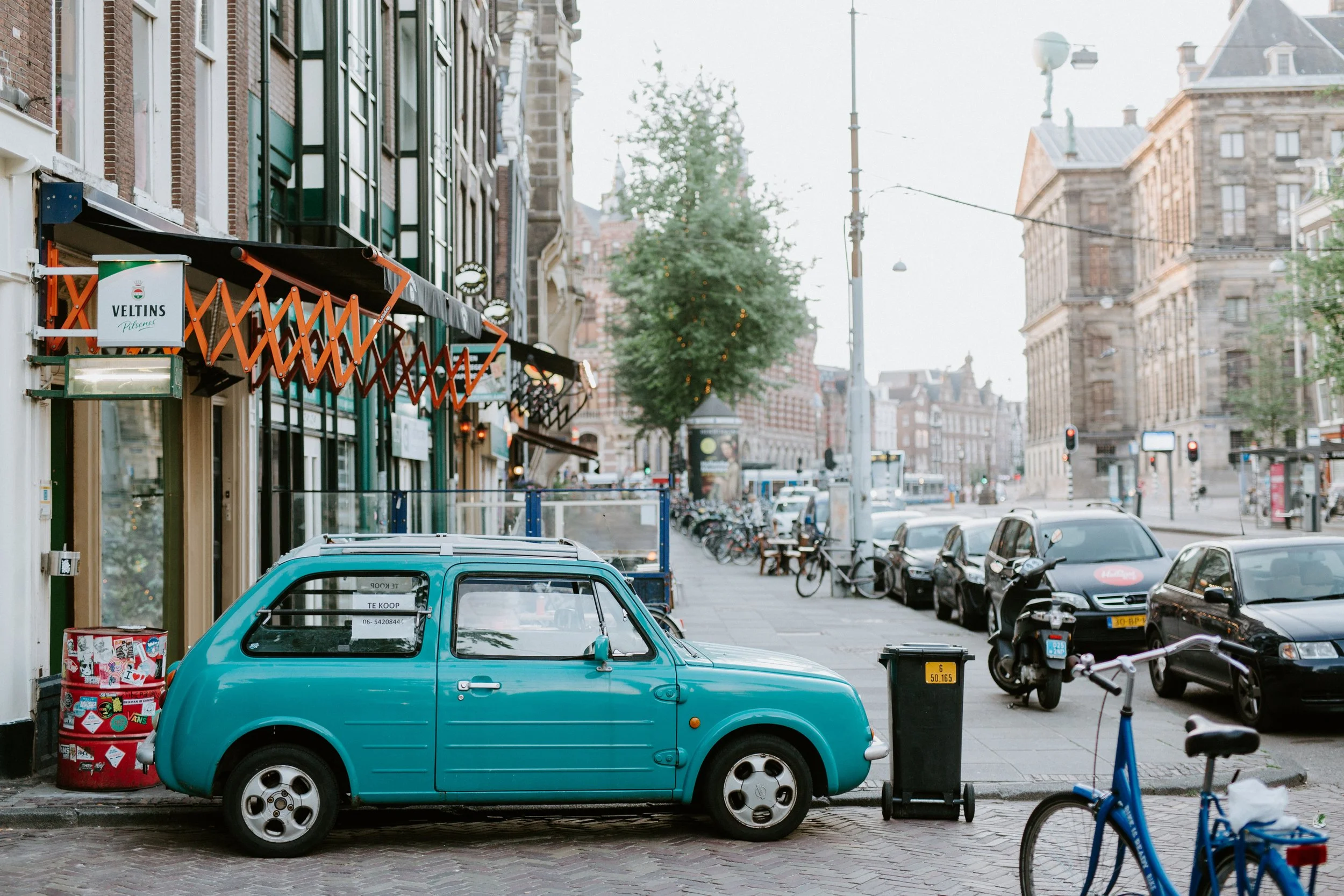 A light blue vintage car parked on a city street sidewalk, next to a trash bin, with bicycles and motorcycles nearby. In the background, there are multi-story buildings, parked cars, and trees along the street, with traffic lights and streetlights vi