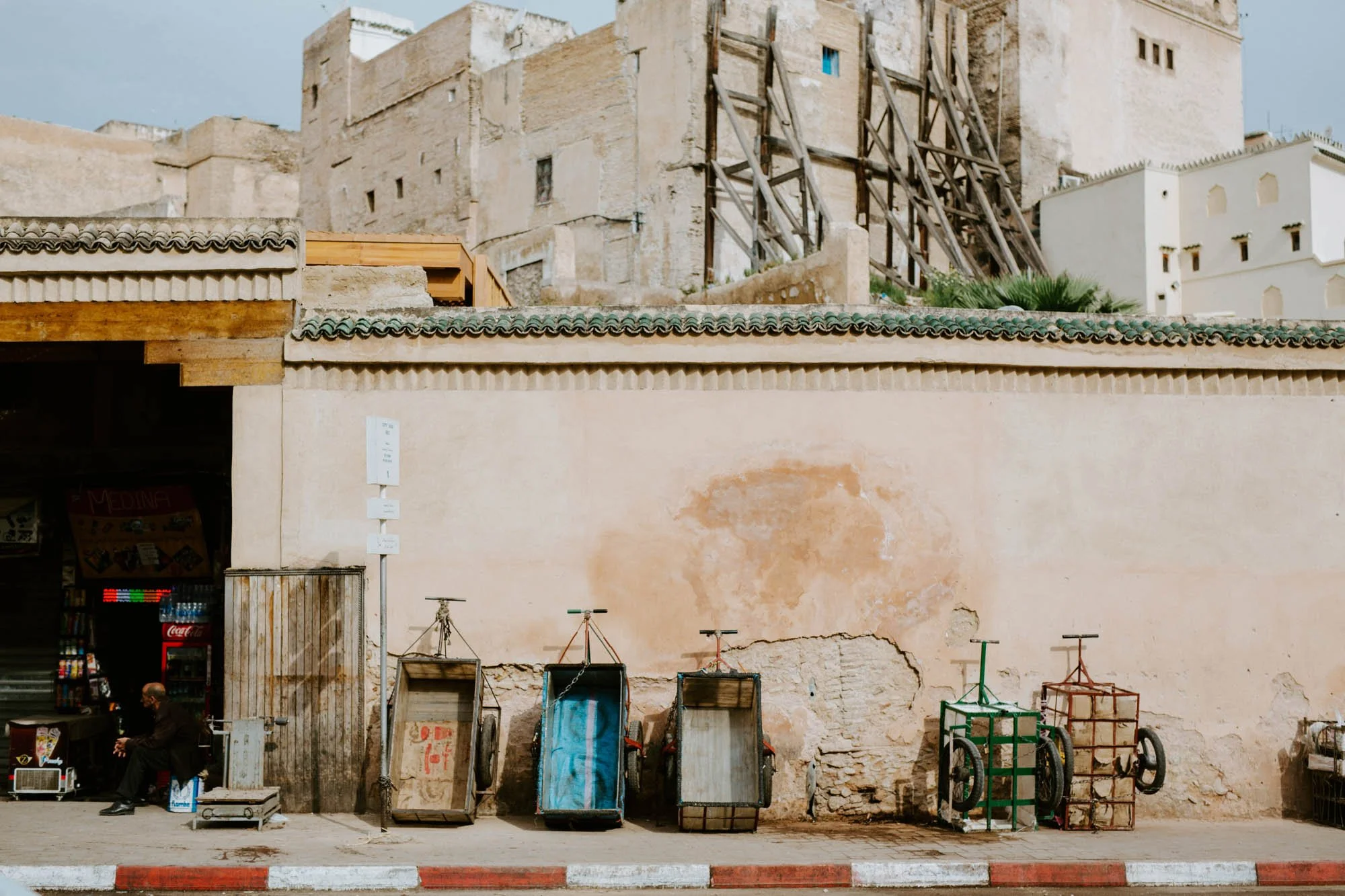 Old, worn building with a cracked wall and six carts in front, some empty and some with items, next to a vending machine and a person sitting on a stool in Fes, Morocco.