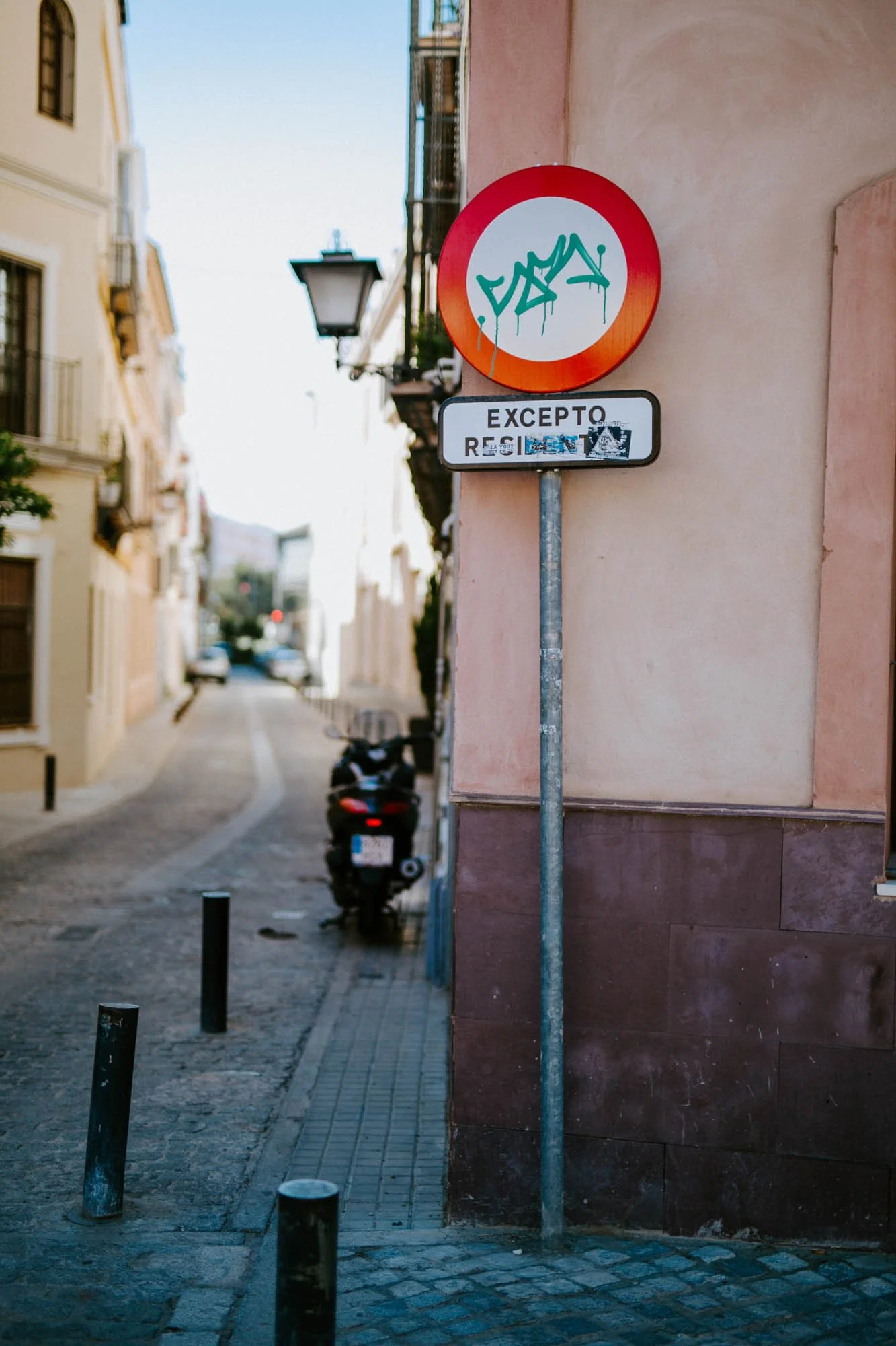 Street sign with graffiti, reading 'EXCEPTO RESIDENTS,' on a narrow street with buildings and a motorcycle.