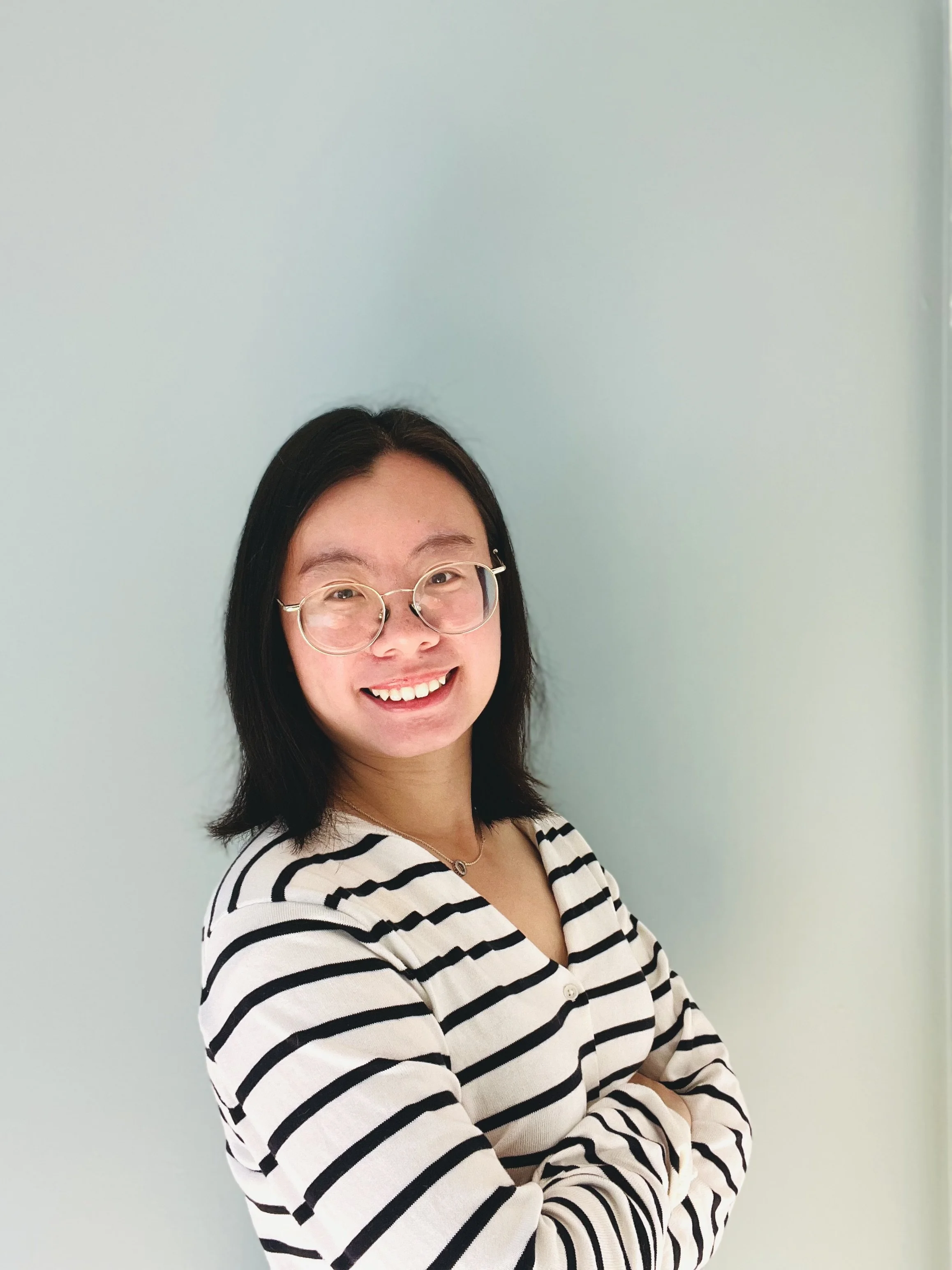 A woman smiling with glasses in front of a plain light-colored wall, wearing a striped shirt.