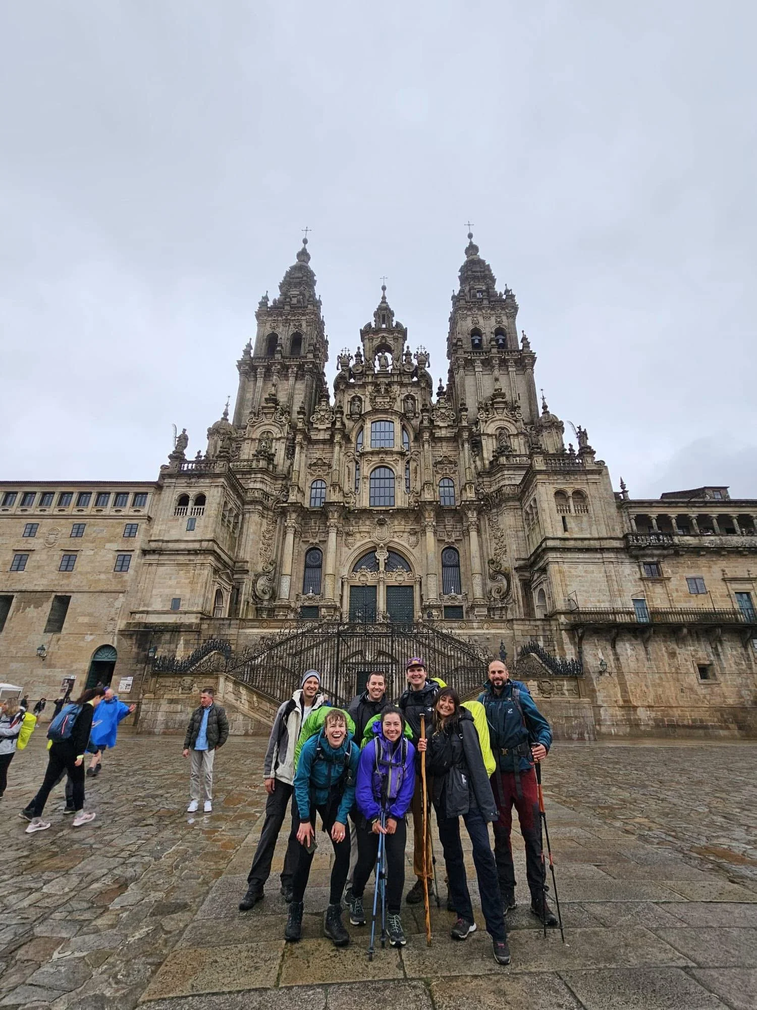 A group of seven hikers standing in front of the Cathedral of Santiago de Compostela, with an overcast sky and wet cobblestone ground.