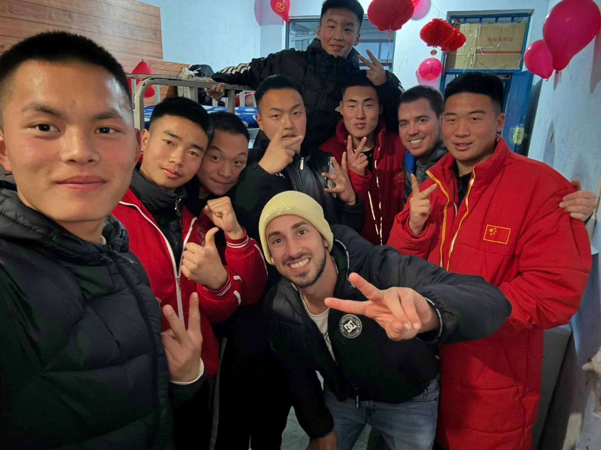 Group of young men, some wearing red jackets with Chinese flag, posing together indoors decorated with red balloons, celebrating.