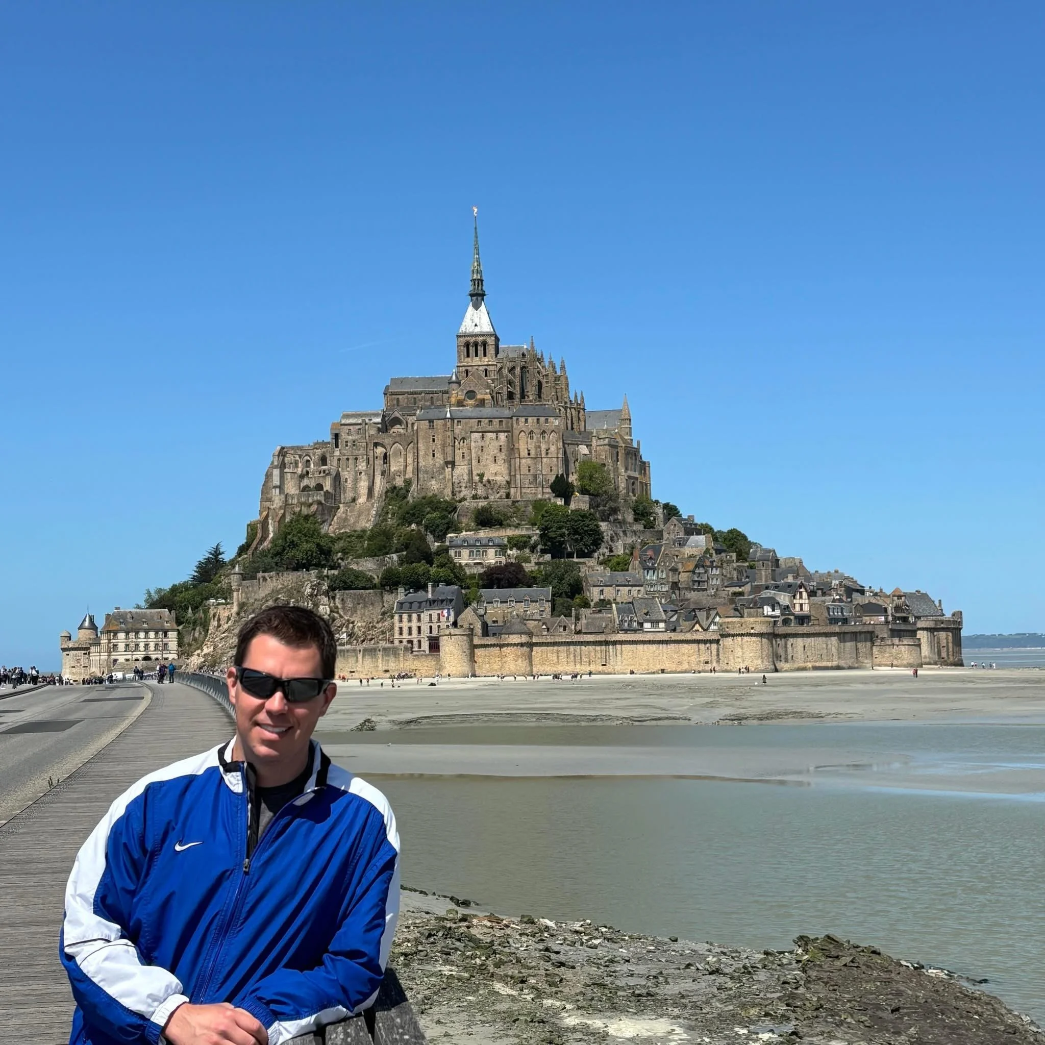 A man in sunglasses and a blue jacket standing in front of Mont Saint-Michel in France with a clear blue sky above.