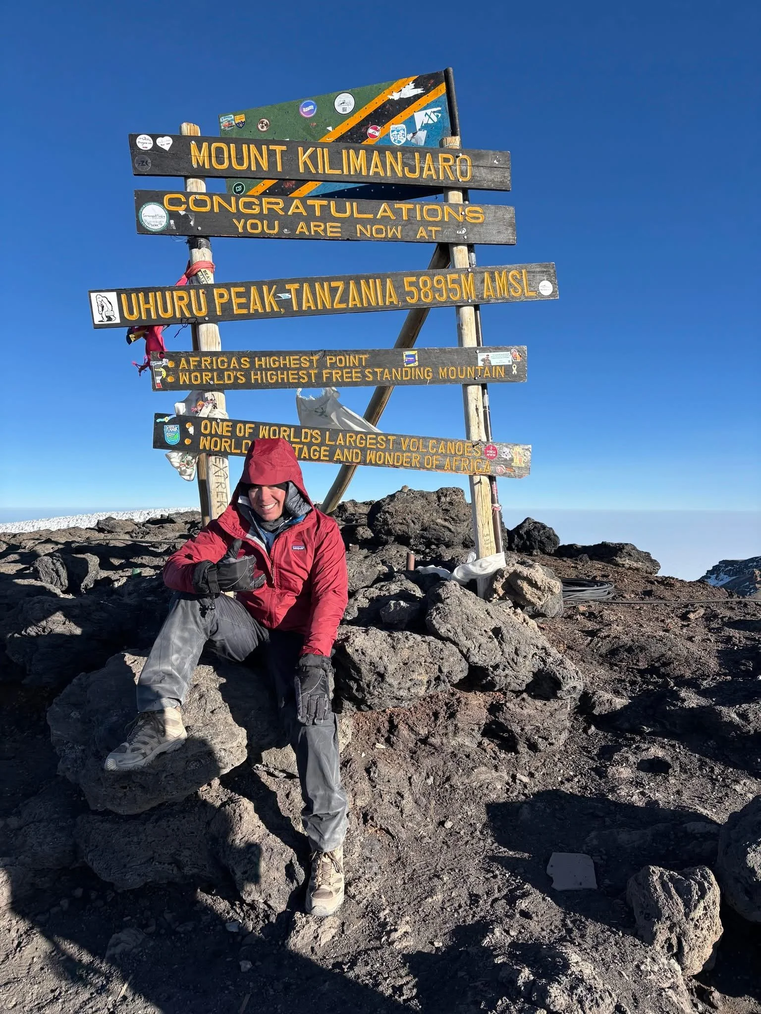 Person in red jacket and gray pants sitting on rocks at summit of Mount Kilimanjaro with wooden signboard indicating the peak at 5895 meters above sea level, Tanzania, Africa.