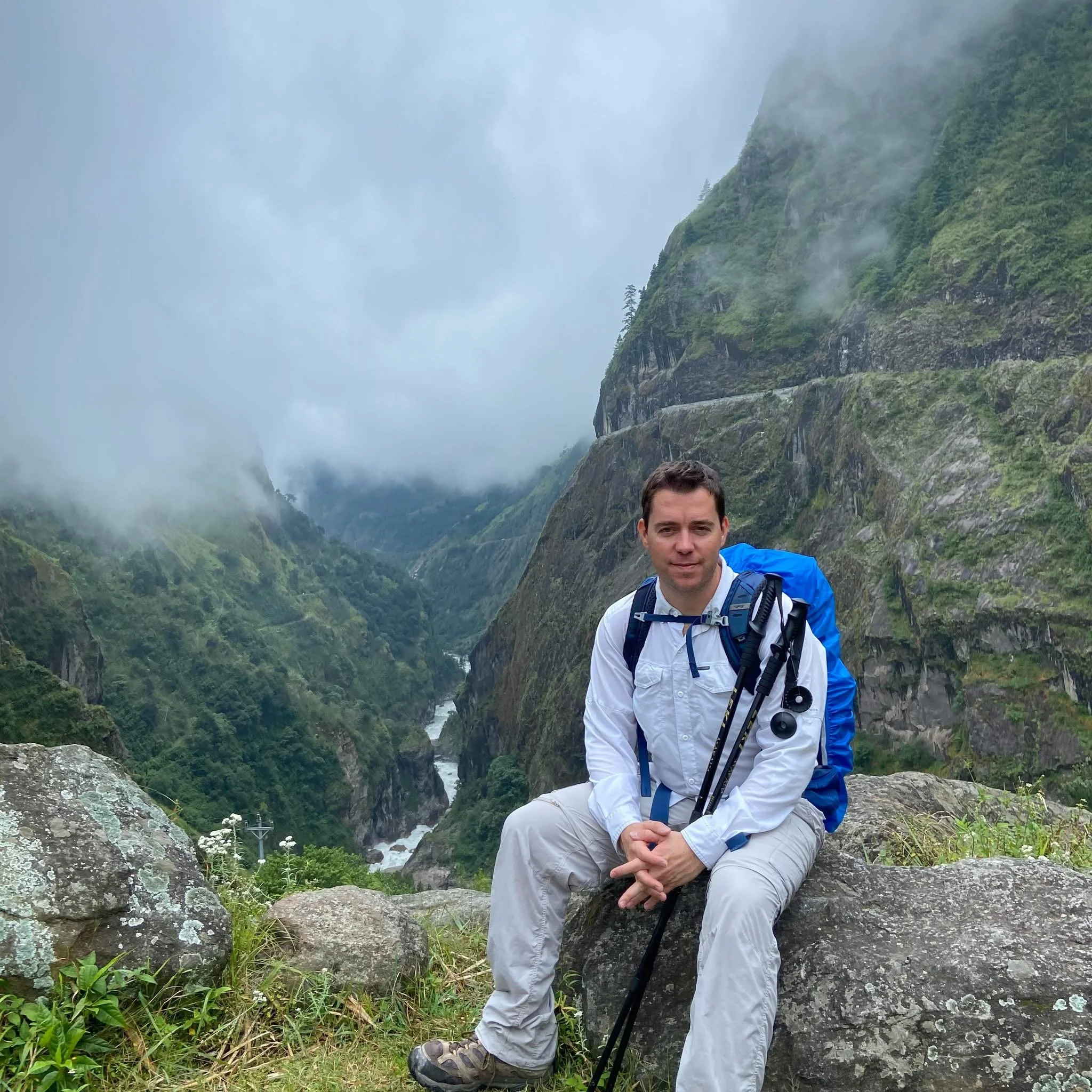 A man sitting on a large rock in a lush, misty mountain valley with a river flowing through it, carrying hiking gear including a blue backpack and hiking poles.