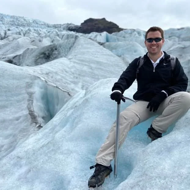 A man in black jacket, beige pants, and sunglasses sitting on a glacier, smiling at the camera, holding a hiking pole, with a backdrop of ice and snow-covered terrain.