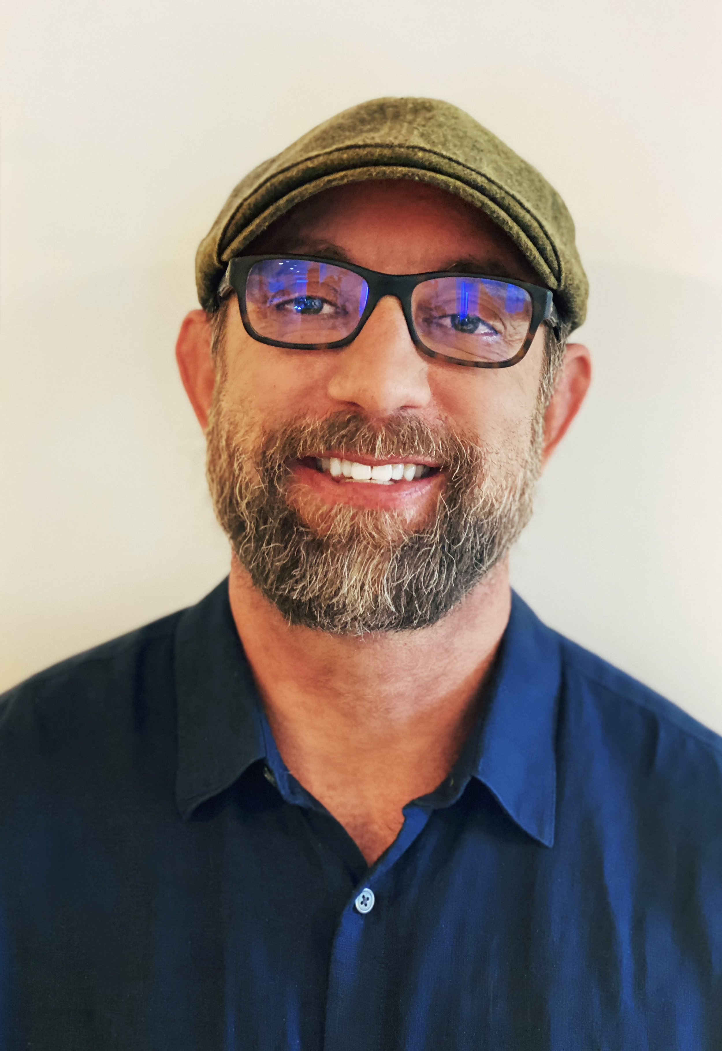 Man wearing glasses, a cap, and a blue shirt, smiling at the camera against a light background.