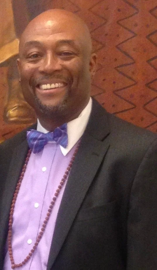 A smiling man dressed in a black suit, lavender shirt, purple bow tie, and wearing a beaded necklace, standing in front of a wooden background with geometric carvings.