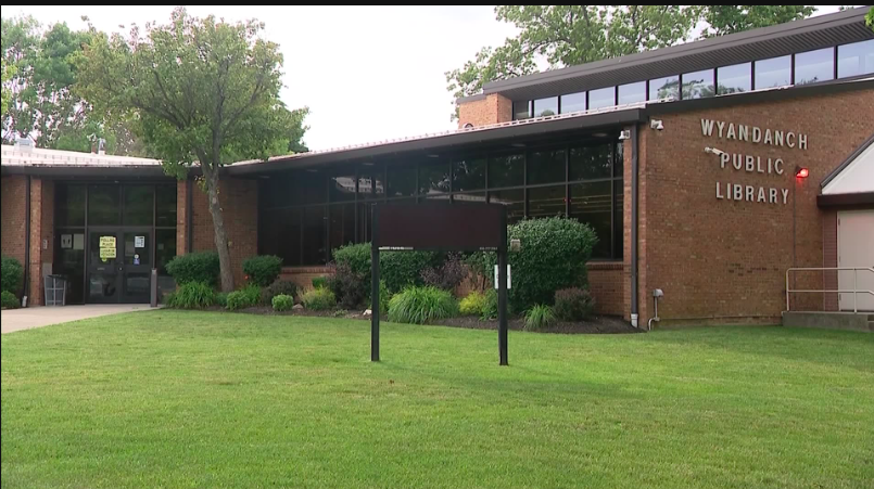 Exterior of Wyandanch Public Library building with grass, bushes, and a large tree, featuring glass windows and signage.