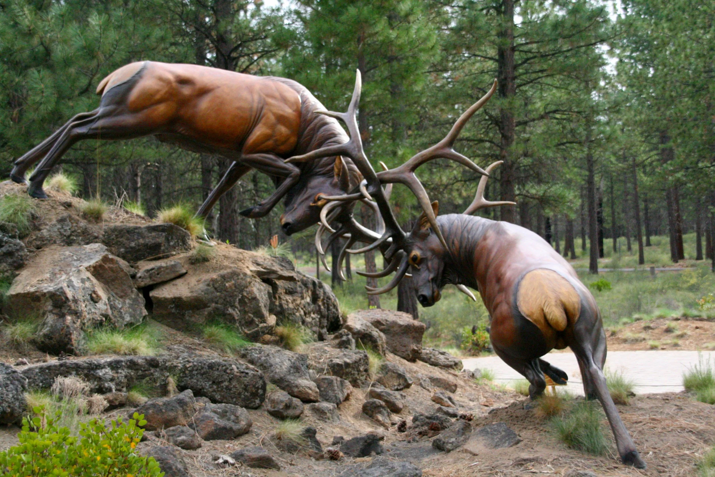 Sculpture of two elk fighting on rocky terrain with trees in the background.