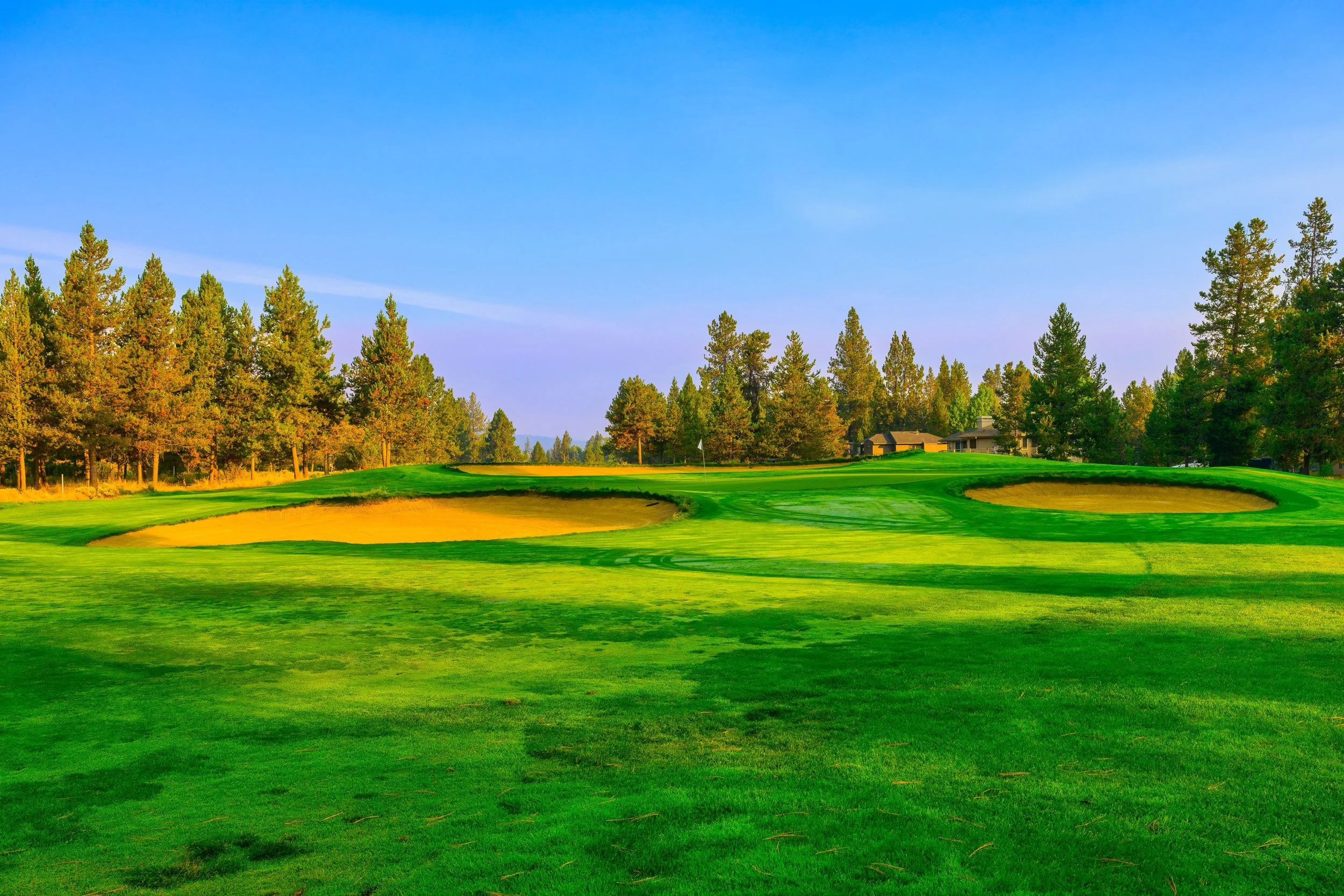 A golf course with green grass, sand bunkers, and a flag on the green, surrounded by trees under a clear blue sky.