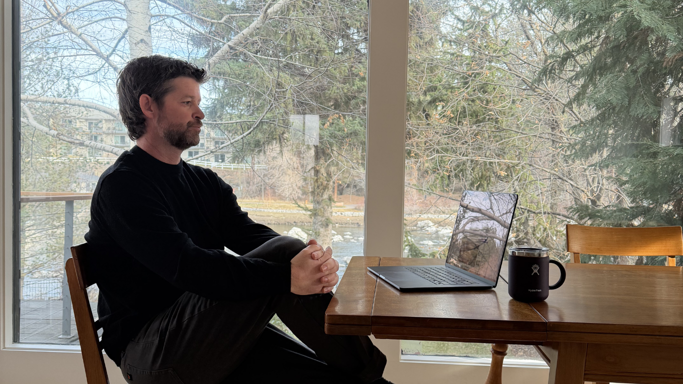 A man sitting at a wooden table in front of large windows, with a laptop and a black mug on the table, overlooking a backyard with trees and a river.