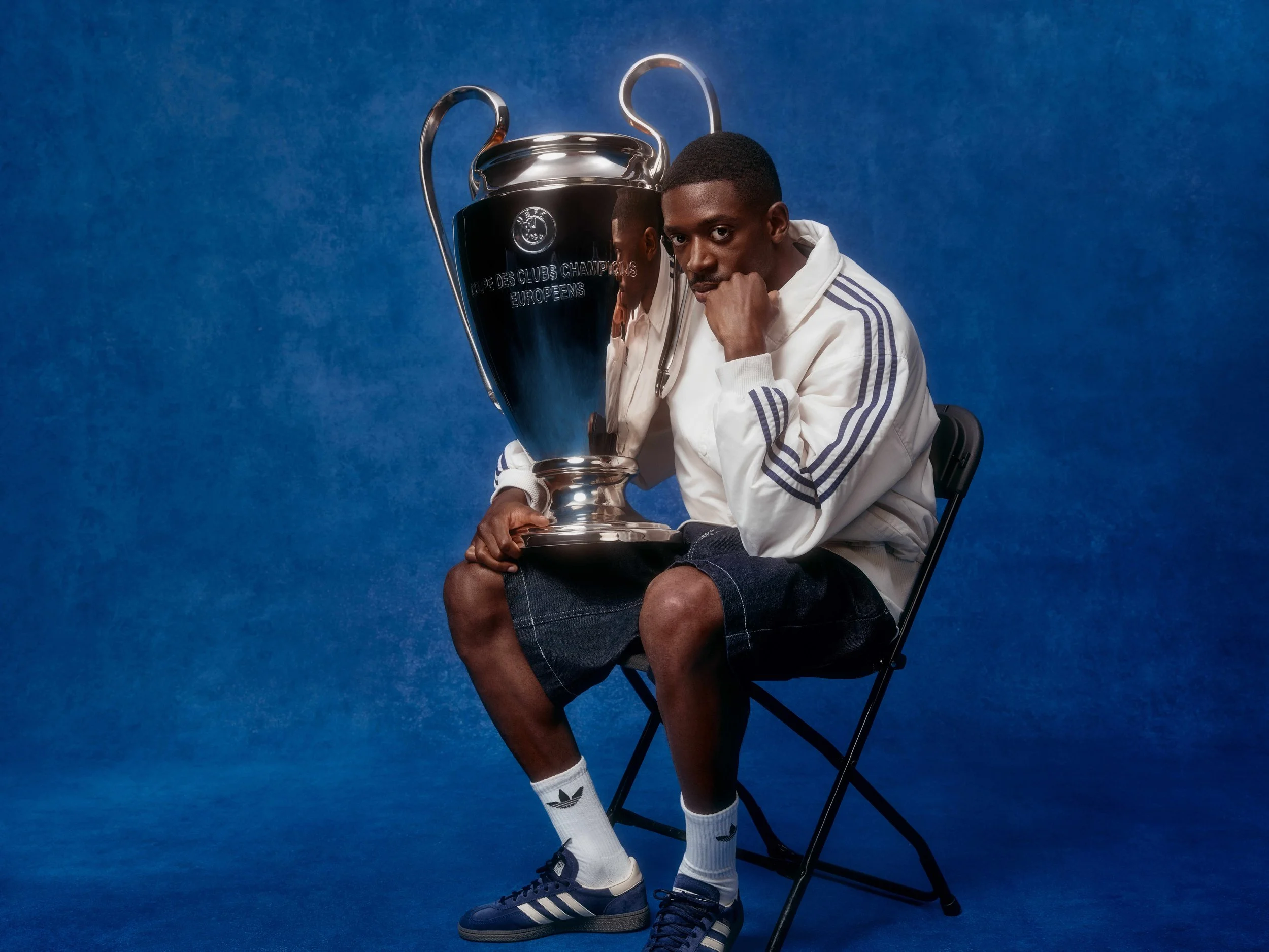 A man sitting on a black folding chair holding a large silver UEFA Champions League trophy. He wears a white Adidas tracksuit jacket with blue stripes, black shorts, white Adidas socks, and blue Adidas sneakers. The background is blue.