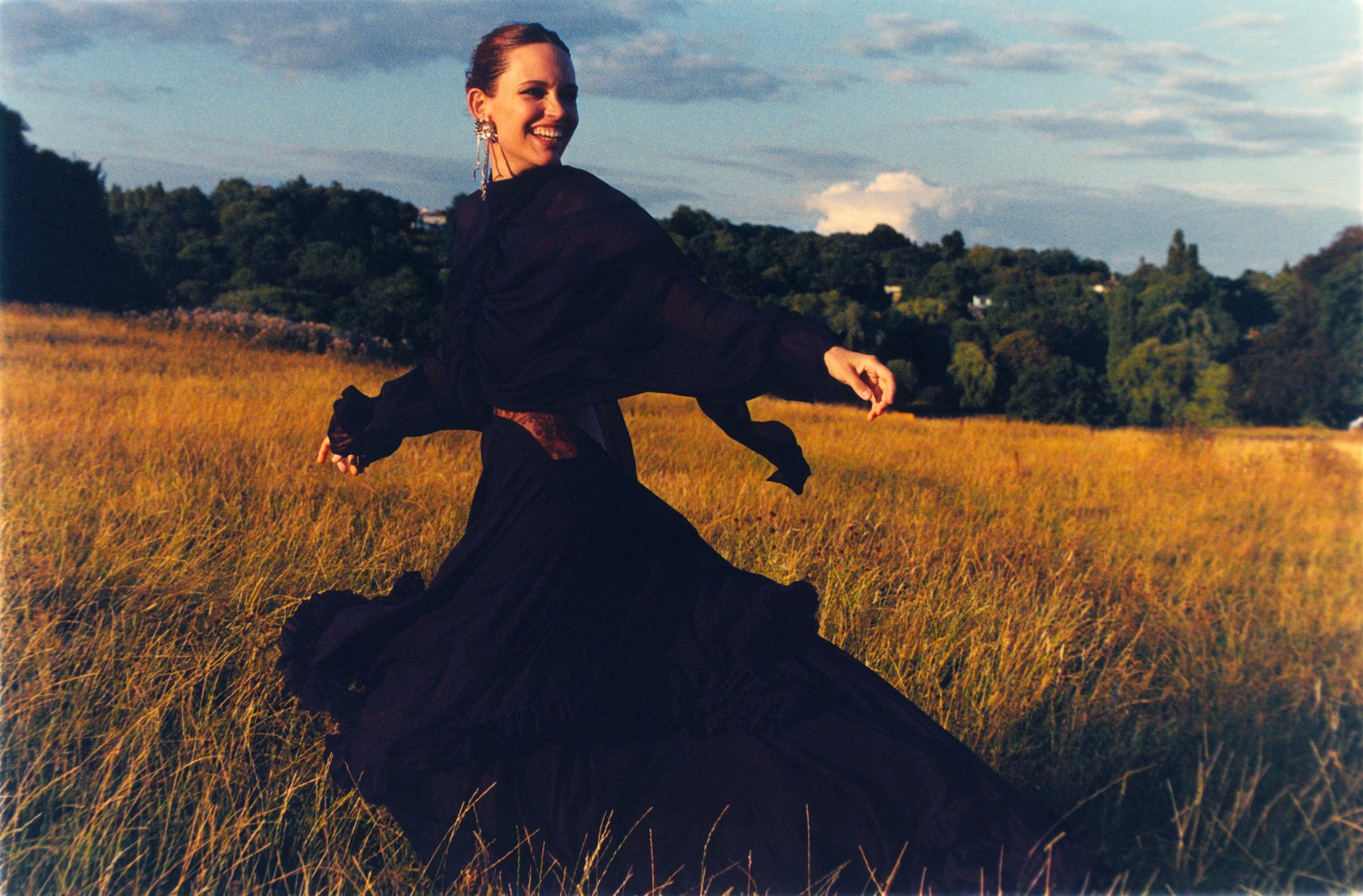 A woman in a flowing black dress twirling in a grassy field with a background of trees and a partly cloudy sky.