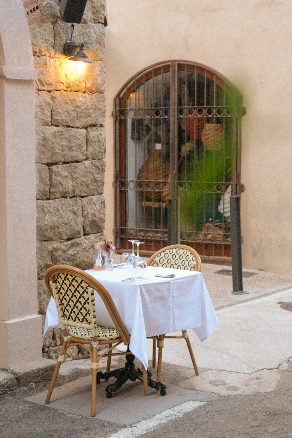 Table de restaurant avec deux chaises, nappes blanches, verres, couverts, dans une rue pavée, derrière une porte en fer forgé.