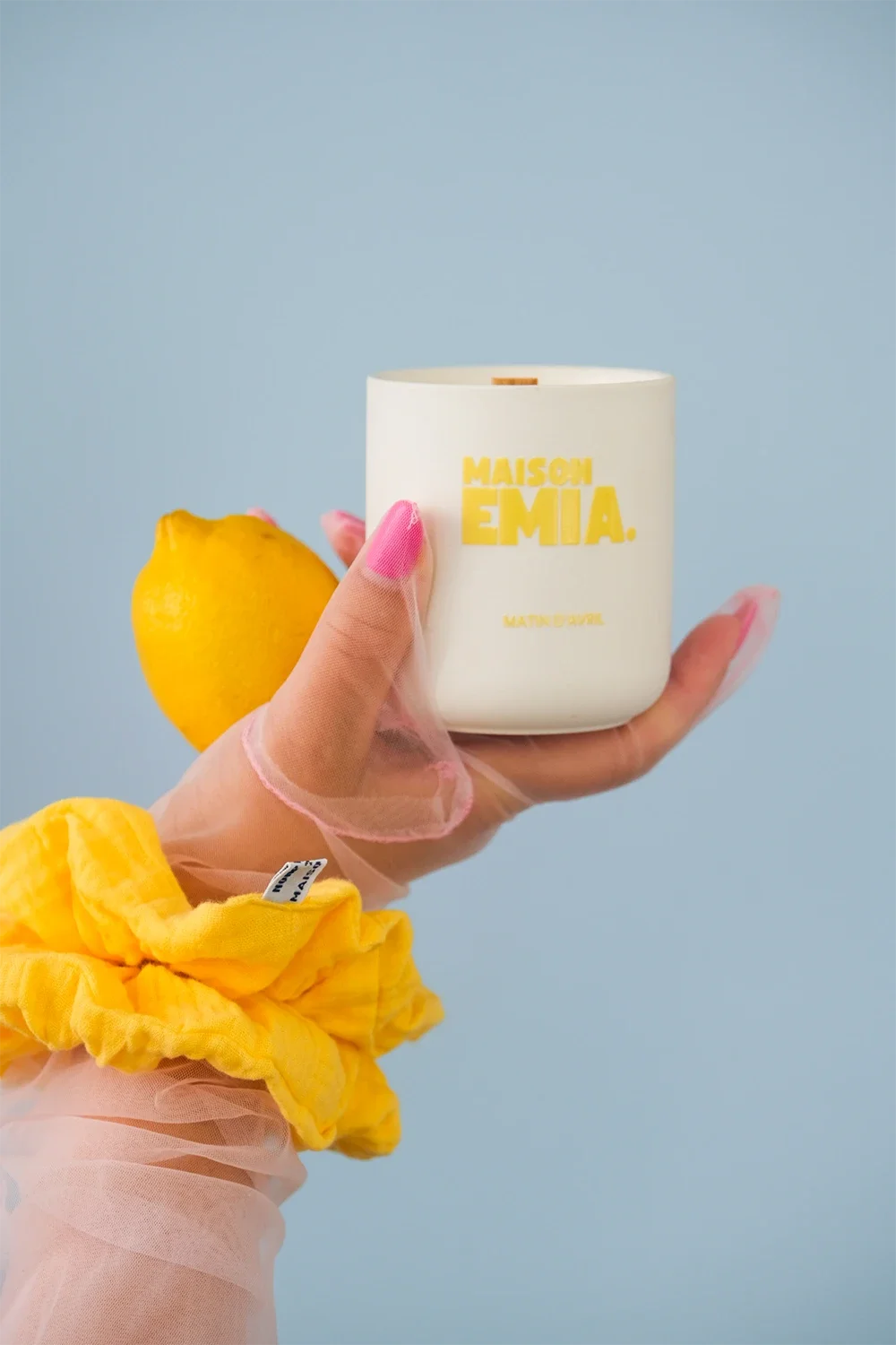 Une main porte un pot blanc avec la marque Maison Emma, une main porte également un citron et une main avec un gant en chiffon jaune. Le fond est bleu clair.
