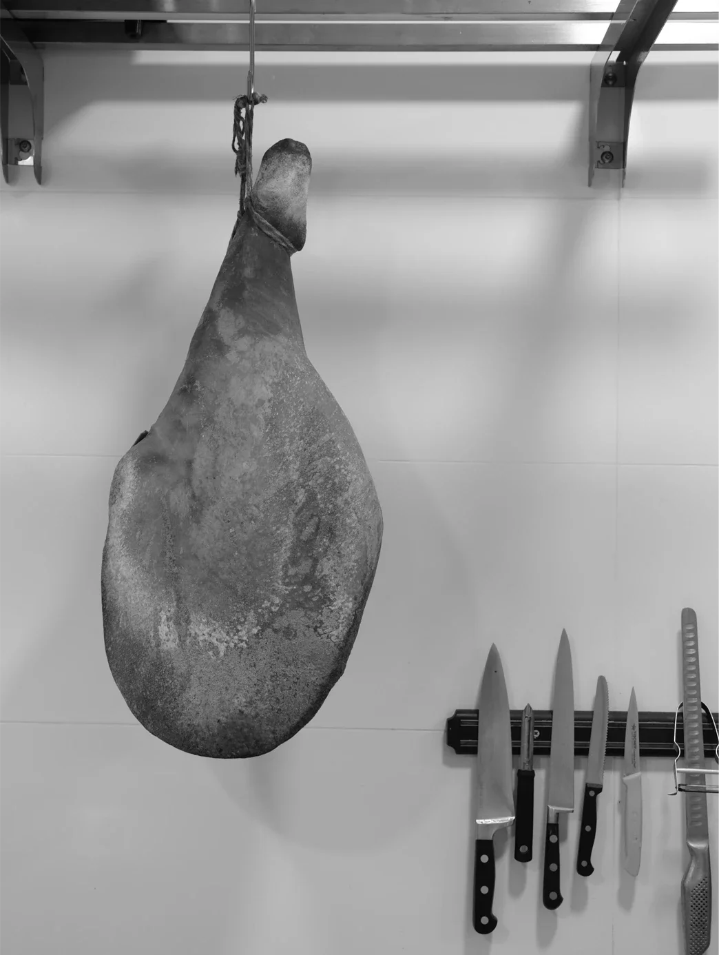 Une pièce de viande suspendue par une corde dans une cuisine, avec plusieurs couteaux de cuisine accrochés au mur en bas à droite.