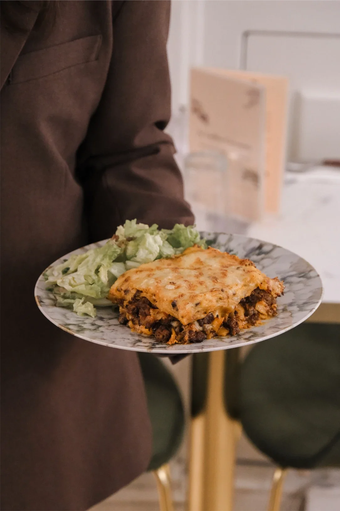 Person tenant un plat de lasagne avec une salade verte à côté dans un cadre intérieur