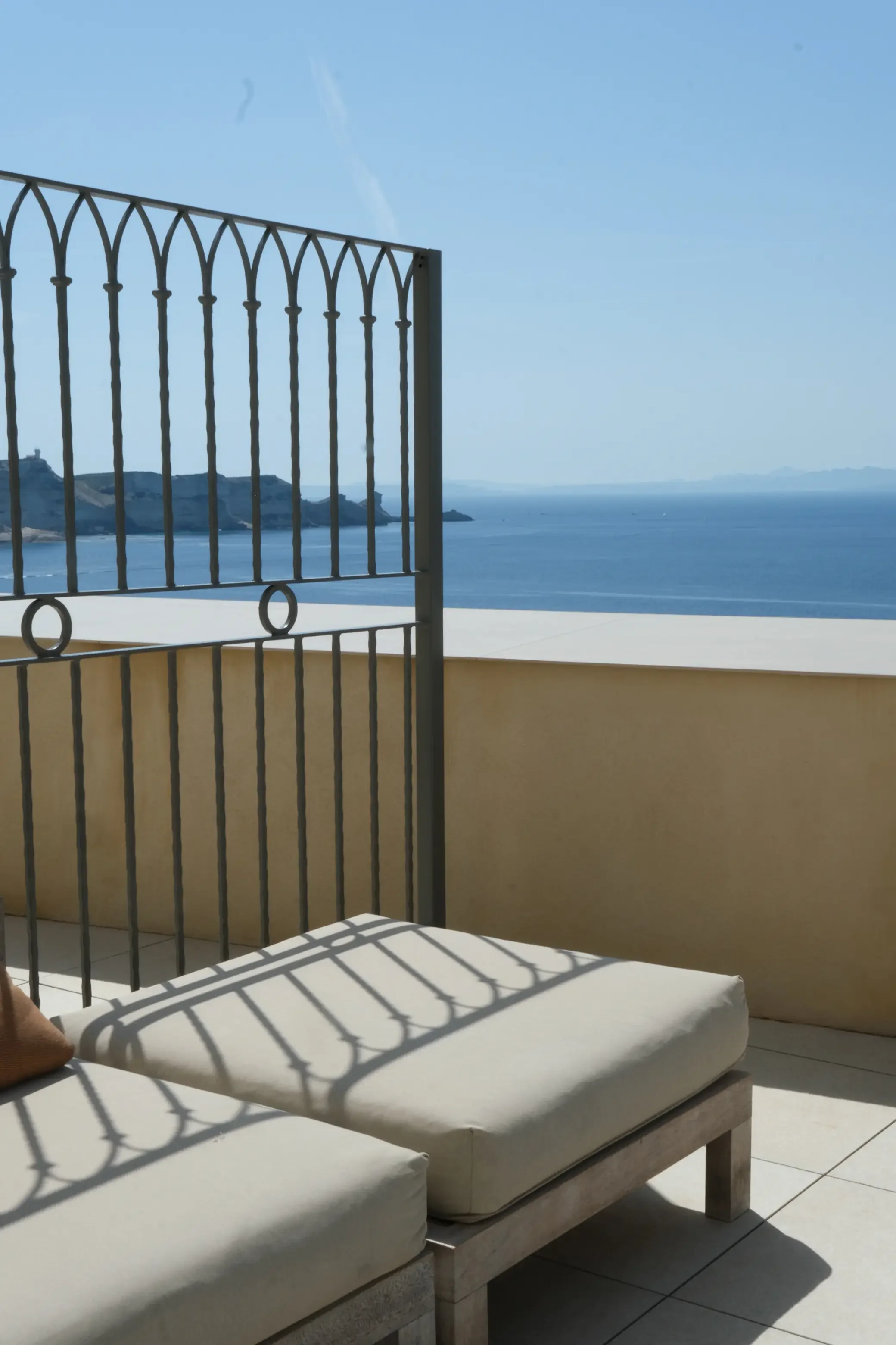 Une terrasse ensoleillée avec deux lits avec des cushions blancs, une barrière en métal avec des arches et des ovales, vue sur la mer avec des falaises au loin, ciel clair et bleu.
