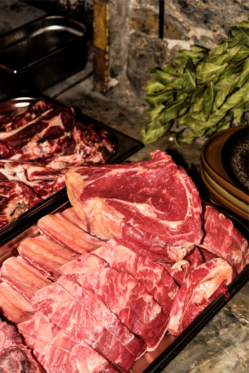 Assortiment de viandes crues, incluant une côte de bœuf et des côtelettes, sur un plateau en métal, avec des herbes vertes en arrière-plan.