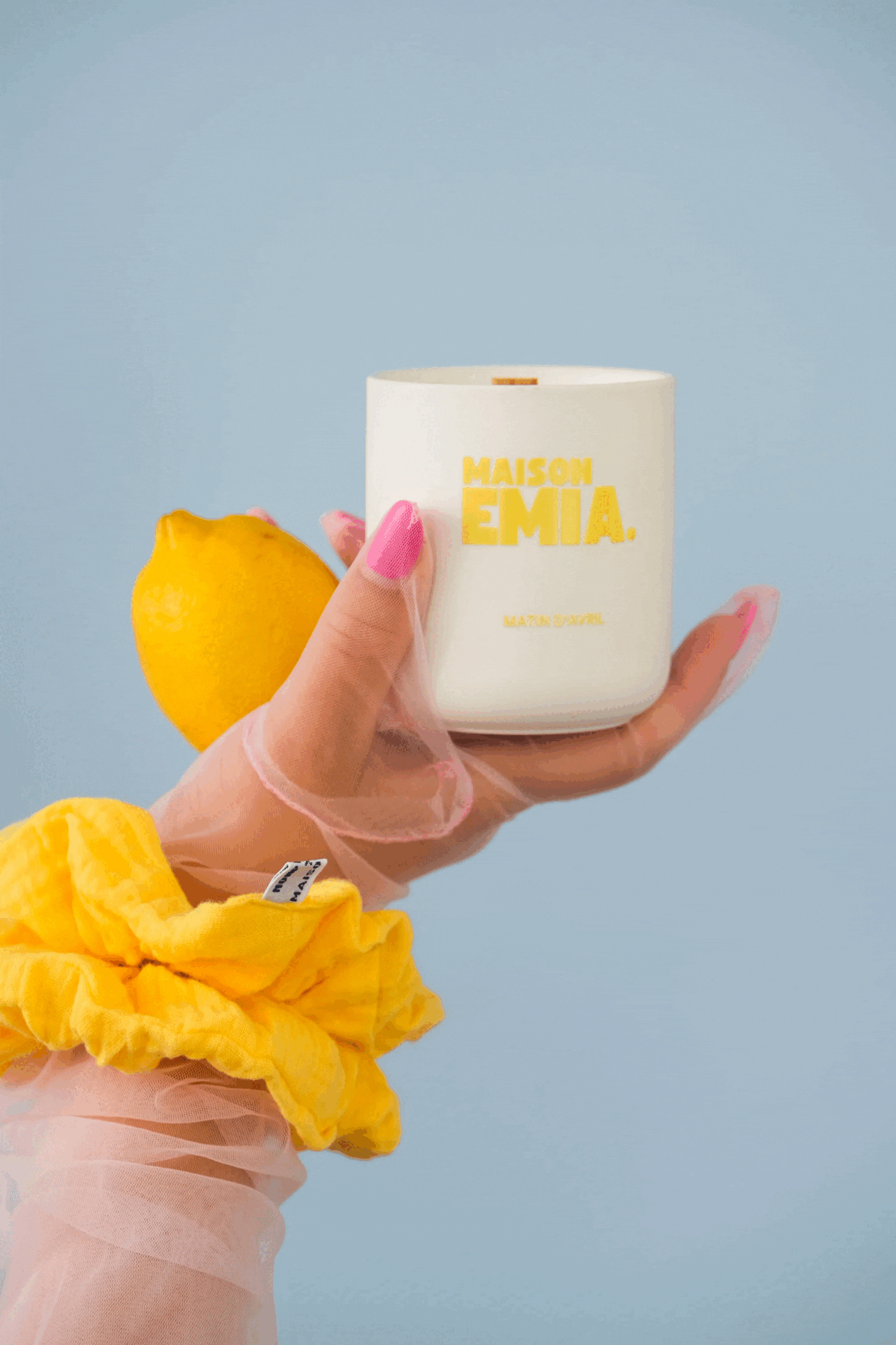 Une main portant un gant en plastique jaune tient un citron, une autre main tient un pot blanc avec le texte "MAISON EMIA" en jaune et un fond bleu clair.