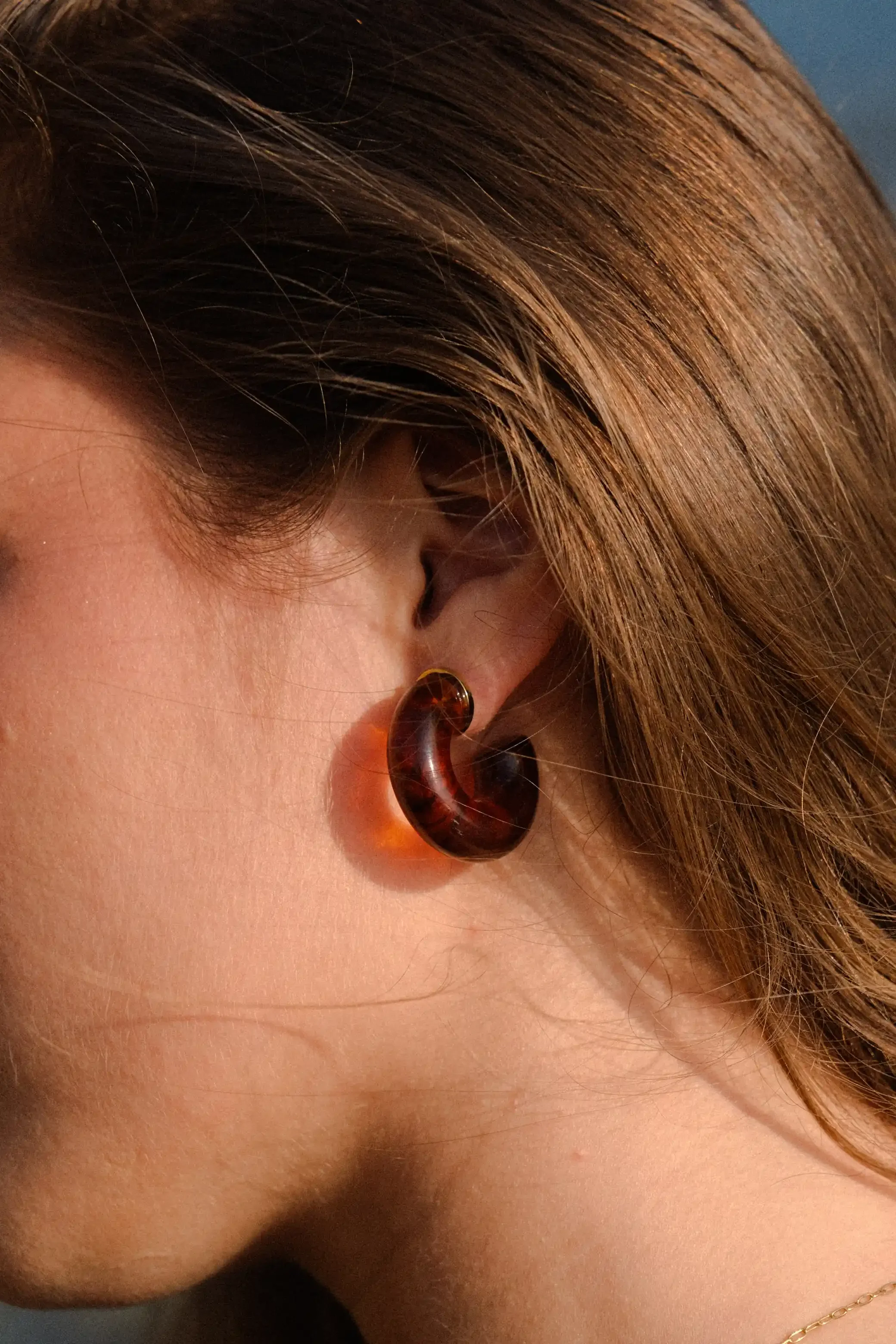 Close-up d'une oreille ornée d'une boucle d'oreille en acétate de couleur marron, avec des cheveux bruns et une peau lisse.