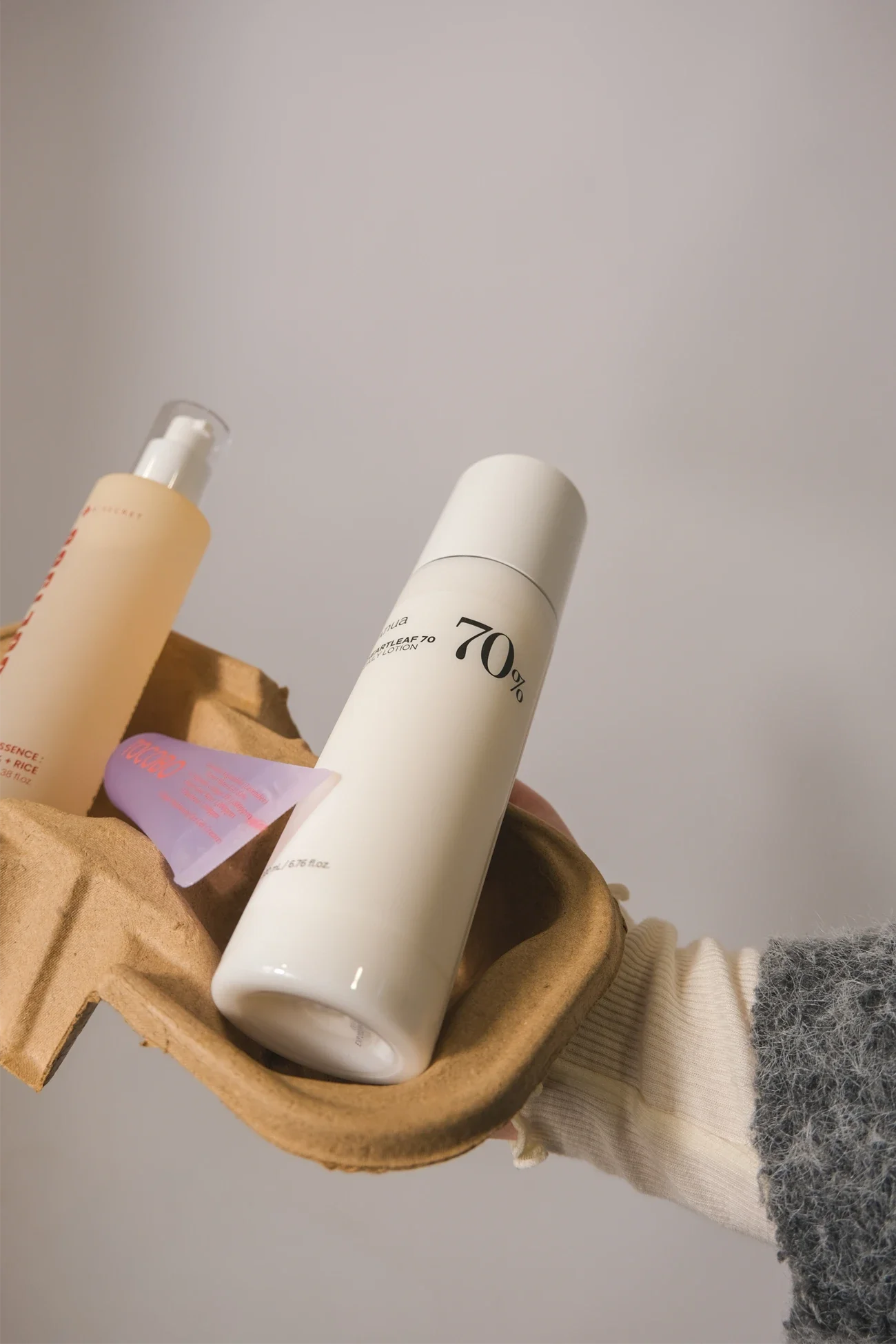 Une main portant un gant en maille grise et un pull beige tient un plateau en carton contenant deux bouteilles de produits cosmétiques ou de soins. L'une est une bouteille blanche avec une pompe, l'autre est une bouteille blanche avec un couvercle. L