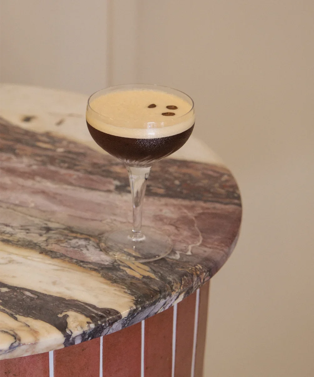 Un verre à cocktail rempli d'un café noir avec une couche de crème chantilly et quelques grains de café sur le dessus, posé sur un comptoir en marbre.