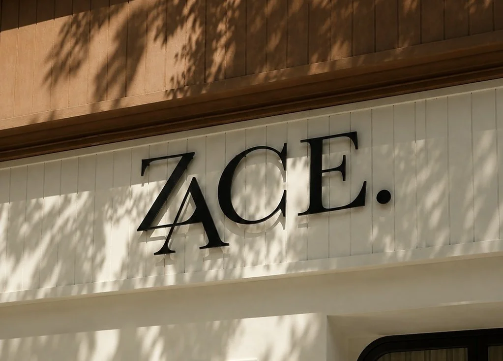 le signe ZAÇE. en lettres majuscules noires sur un fond blanc, avec des ombres de feuilles d'arbre projetées dessus.