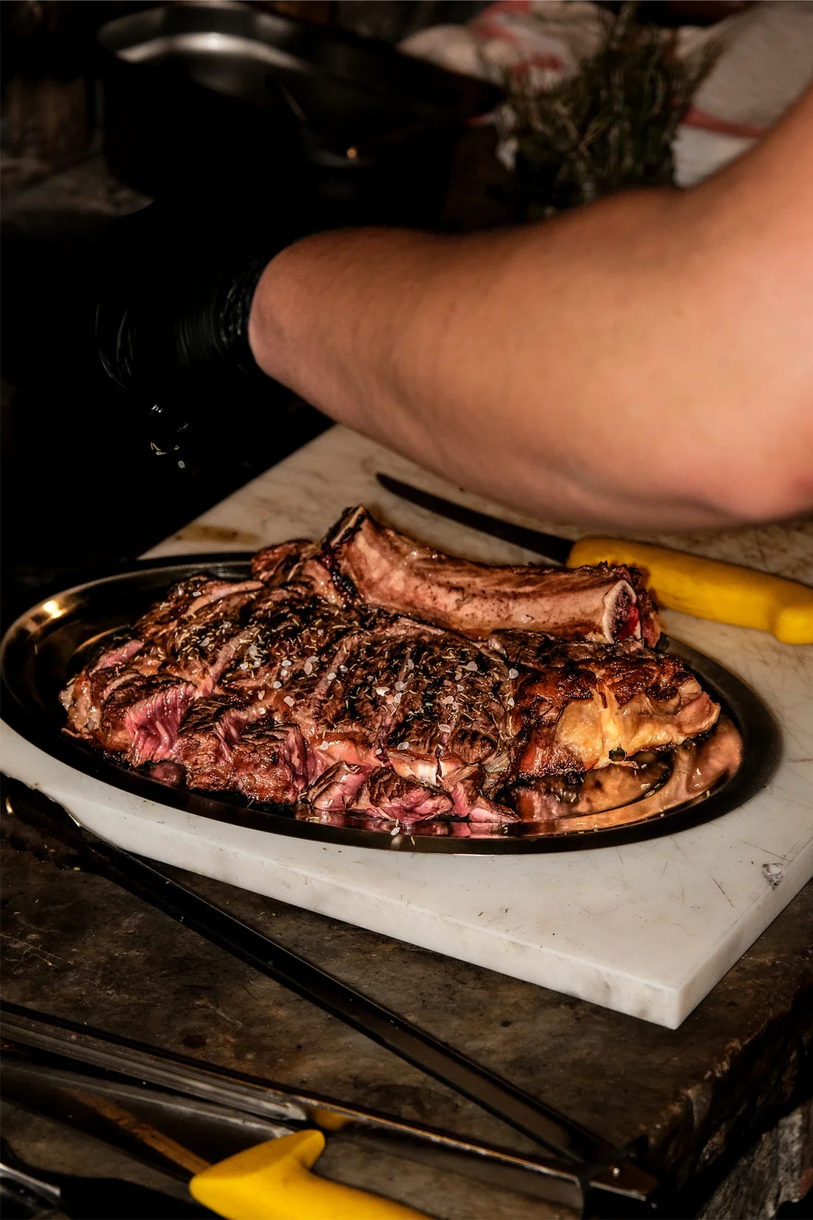 Une pièce de viande de bœuf grillée, assaisonnée de sel, posée sur une assiette en métal noire, sur une surface en marbre. Au fond, un bras d'une personne portant un gant noir, utilisant une pince pour manipuler la viande, accompagné d'un poivron jau