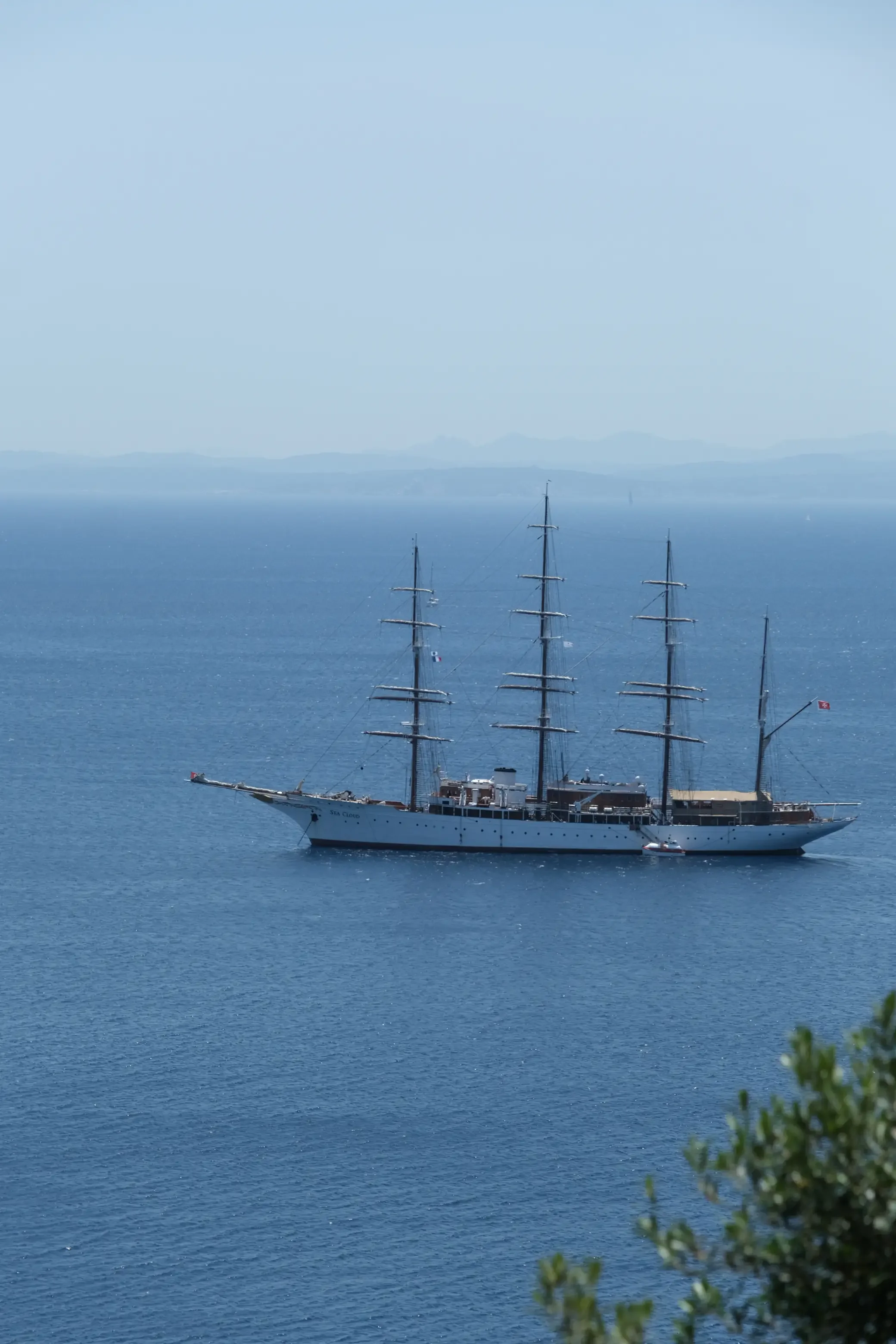 Un grand voilier blanc à plusieurs mâts navigue sur une mer calme, avec des montagnes visibles à l'arrière-plan et un peu de végétation au premier plan.