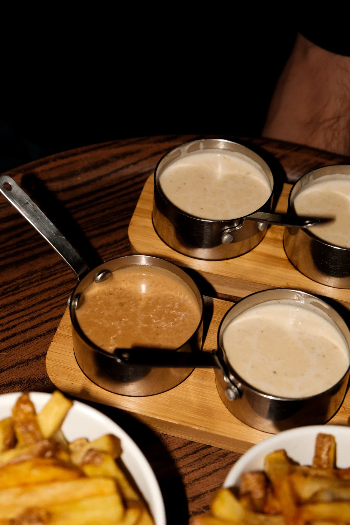 Différents types de sauces dans des petits pots en acier inoxydable, placés sur des plateaux en bois, accompagnant des frites dans des bols.