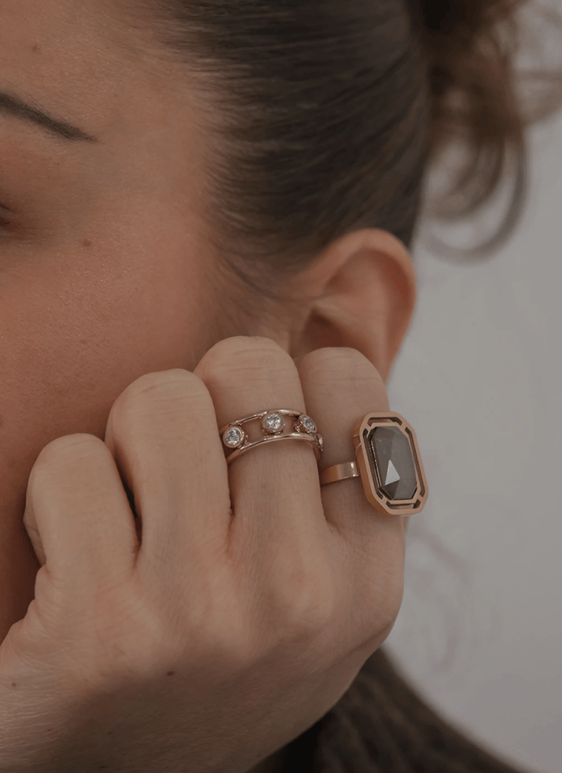 Main avec deux bagues portant des pierres précieuses, près de l'oreille d'une femme aux cheveux bruns.
