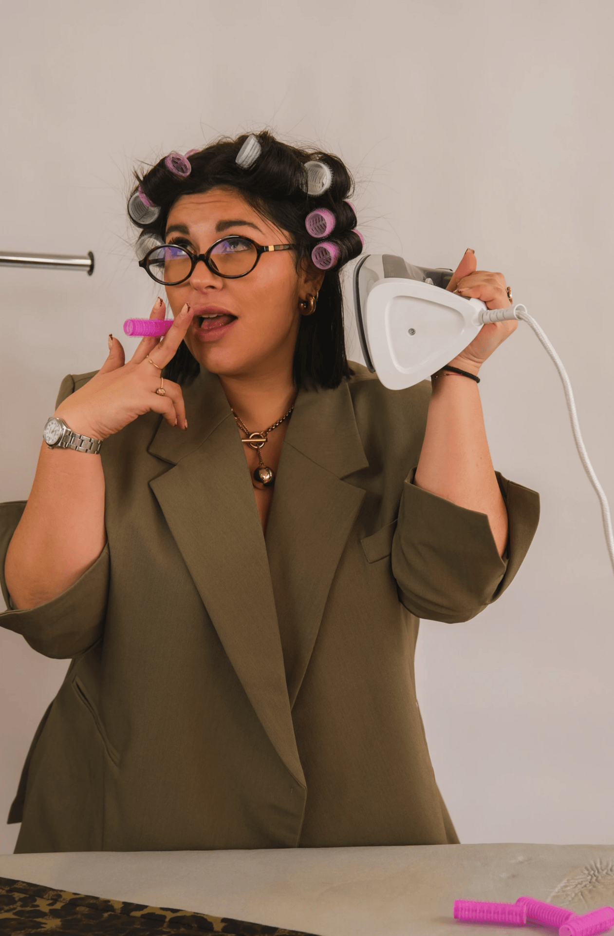 Une femme portant des rouleaux de cheveux, des lunettes et un tailleur beige, pose en tenant un fer à repasser près de sa tête, avec des rouleaux roses sur une table.