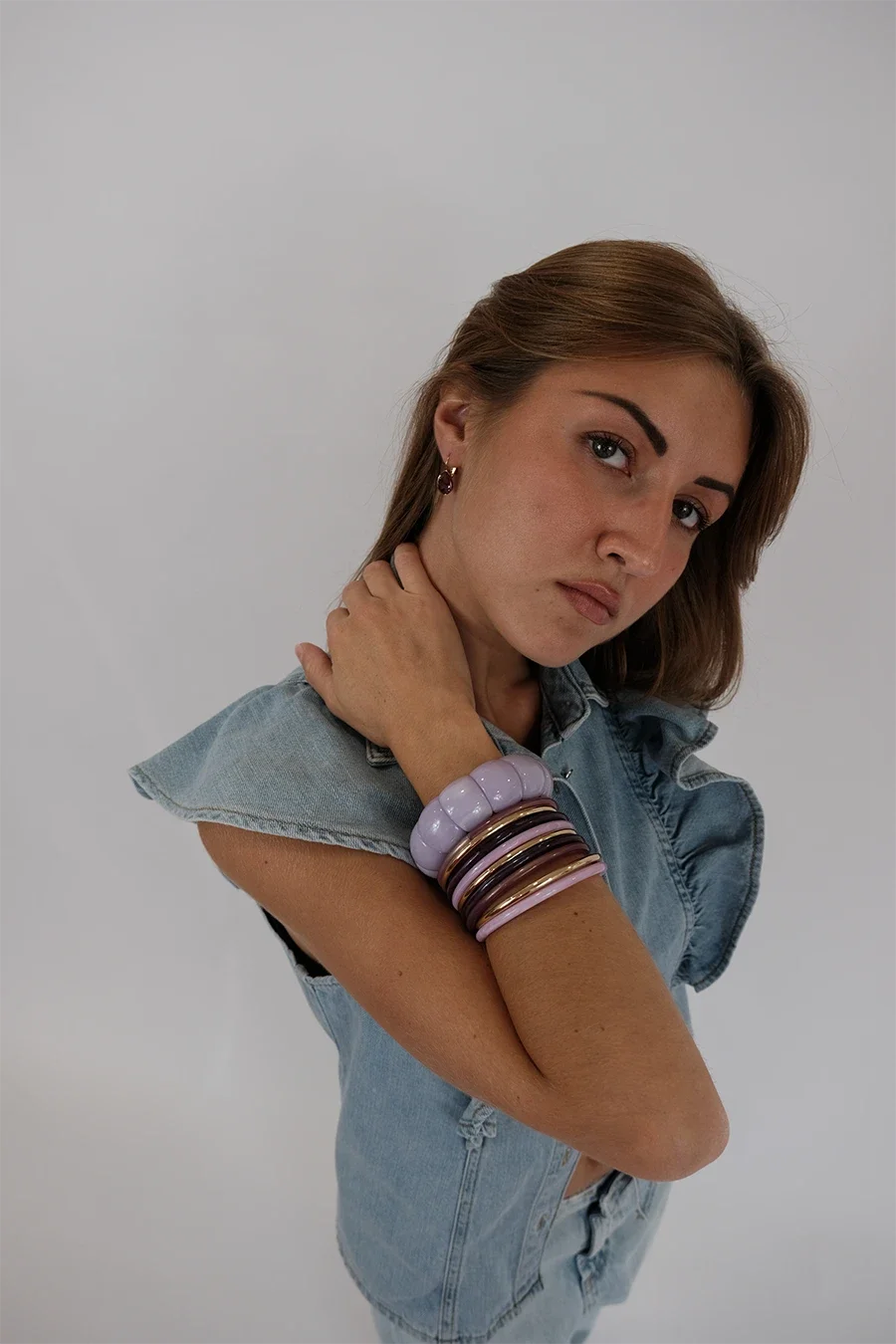 Jeune femme avec bracelets violets et marron, porte un jean déchire et une veste en jean, pose contre un fond blanc.