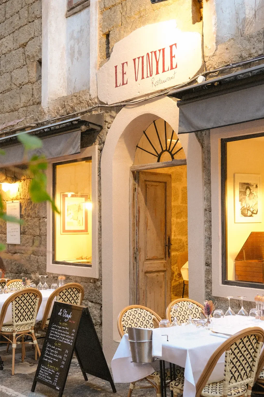 Façade d'un restaurant appelé 'Le Vinyle', avec des fenêtres éclairées, une porte en bois, des chaises en rotin avec un tapis de nappe blanche, des tables avec verres et un menu affiché en extérieur.