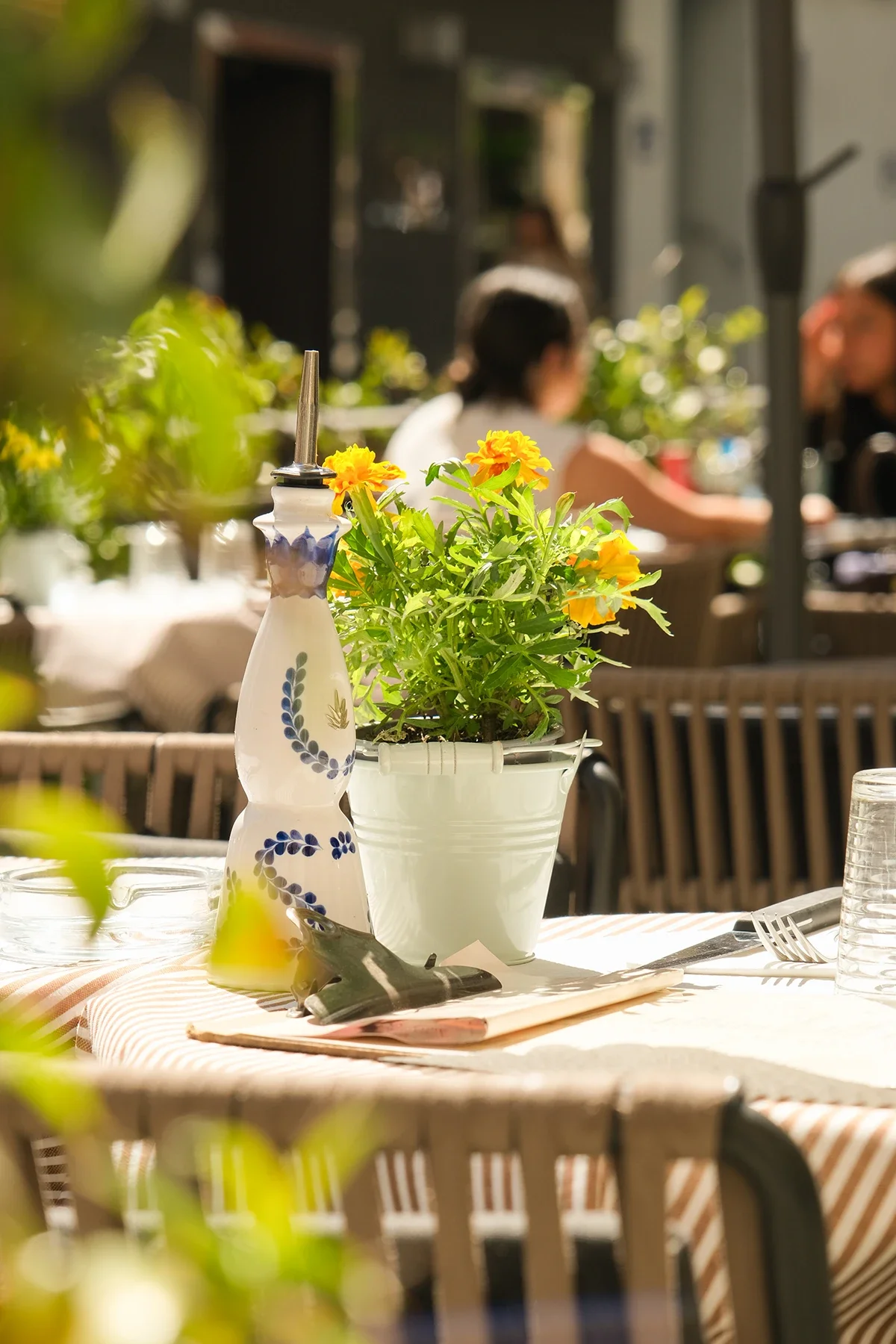 Une table de restaurant en plein air avec un vase blanc contenant des fleurs jaunes et une bouteille en céramique décorative. En arrière-plan, deux personnes assises parlant, floues. La scène est baignée de lumière naturelle.