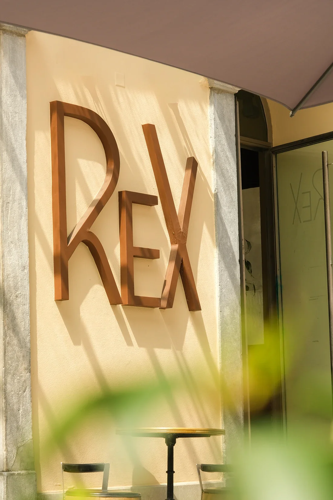 Une enseigne en métal formant le mot "REX" fixée sur un mur beige. La scène est en extérieur, avec un parasol ou ombrelle en haut à droite et des chaises et une petite table au premier plan.