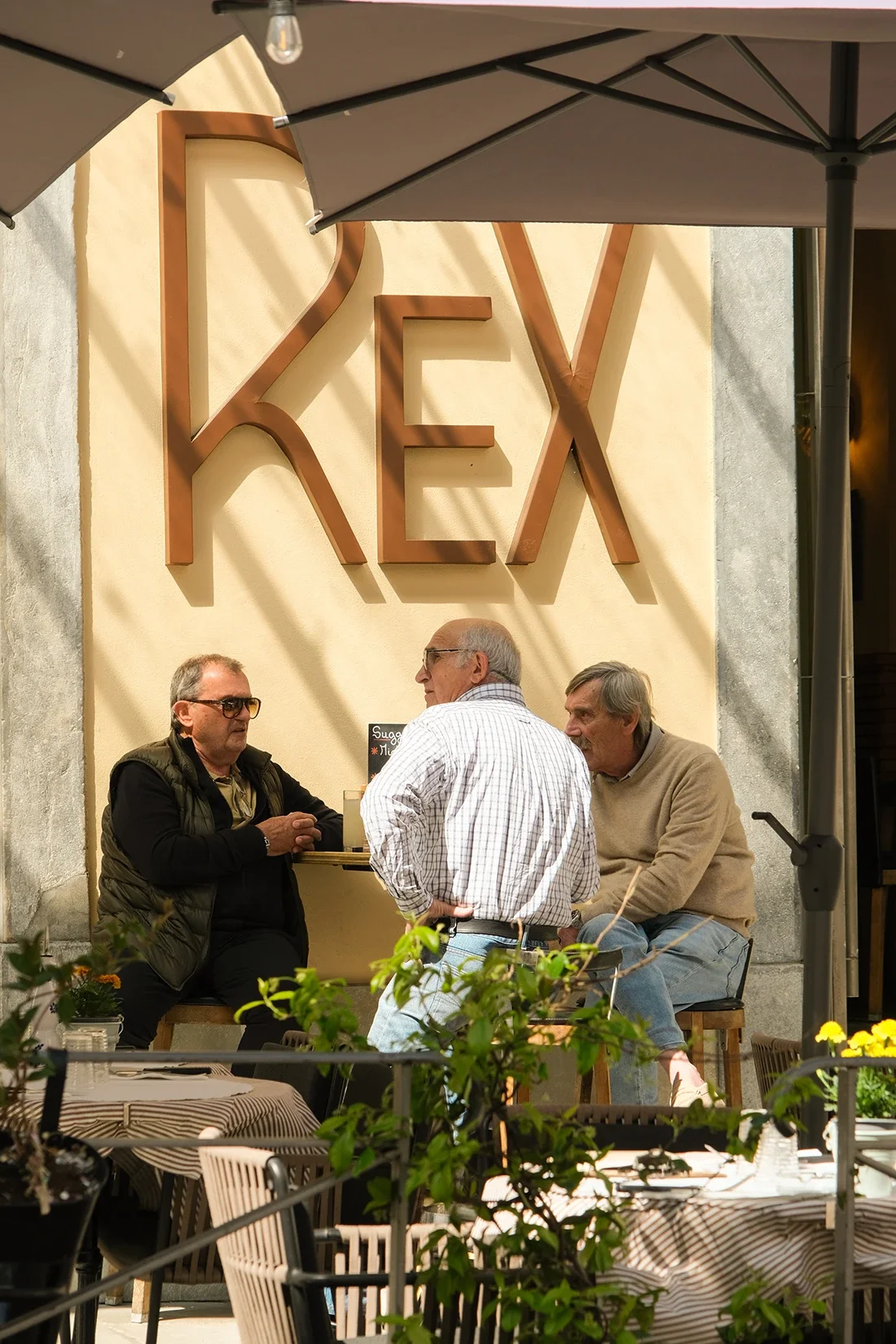 Trois personnes discutent en extérieur devant une façade avec un grand signe « REX ». Un parasol blanc est au-dessus d'eux et des tables avec des plantes en premier plan.